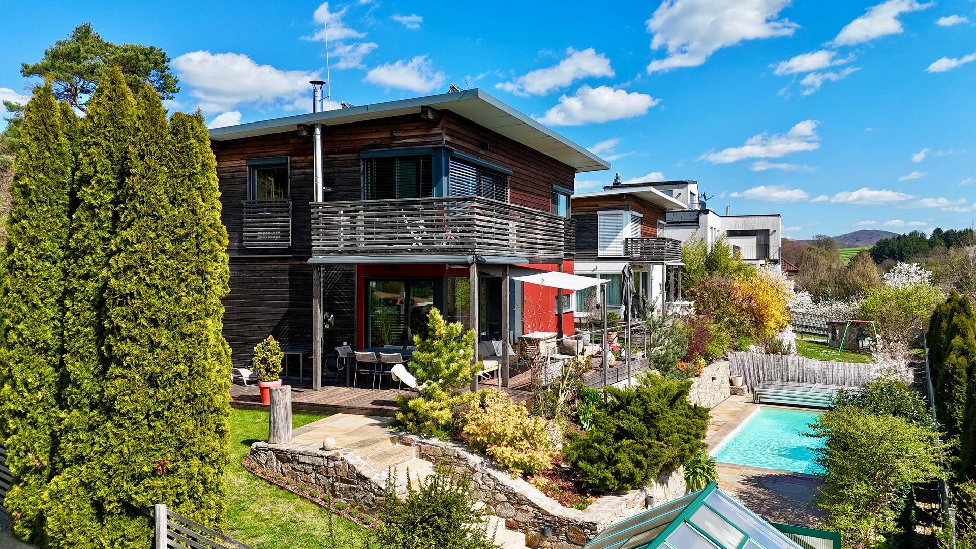 Modernes Haus mit Holzfassade, großem Garten, Terrasse und Swimmingpool unter blauem Himmel.