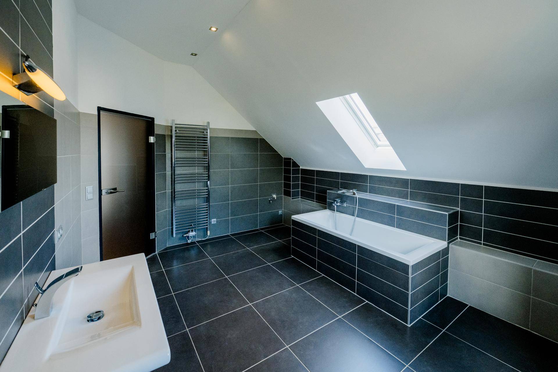 Modernes Badezimmer mit dunklen Fliesen, Badewanne, Waschbecken und beheiztem Handtuchhalter.