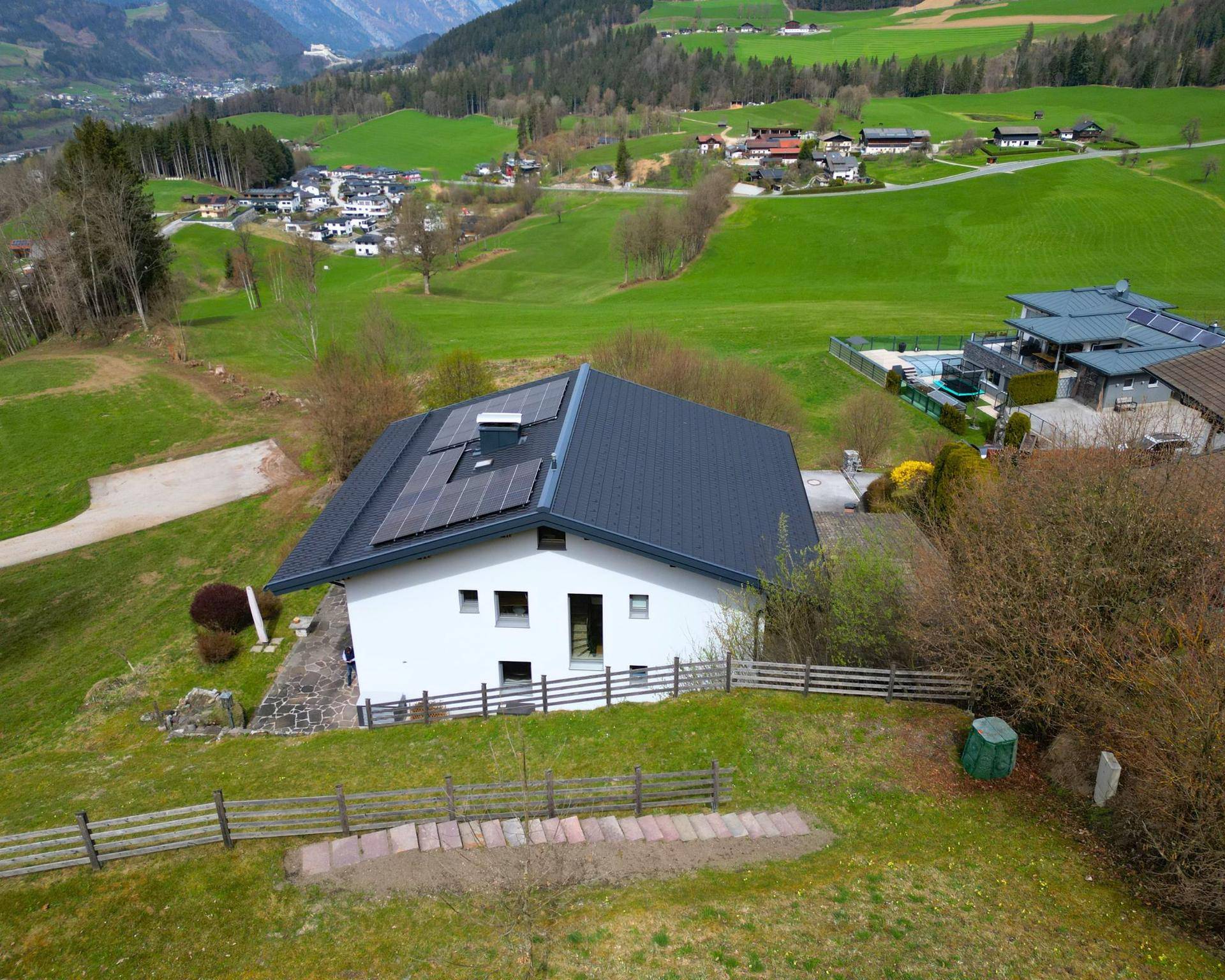 Luftaufnahme eines modernen Hauses mit Solaranlage auf dem Dach, umgeben von grünen Hügeln.