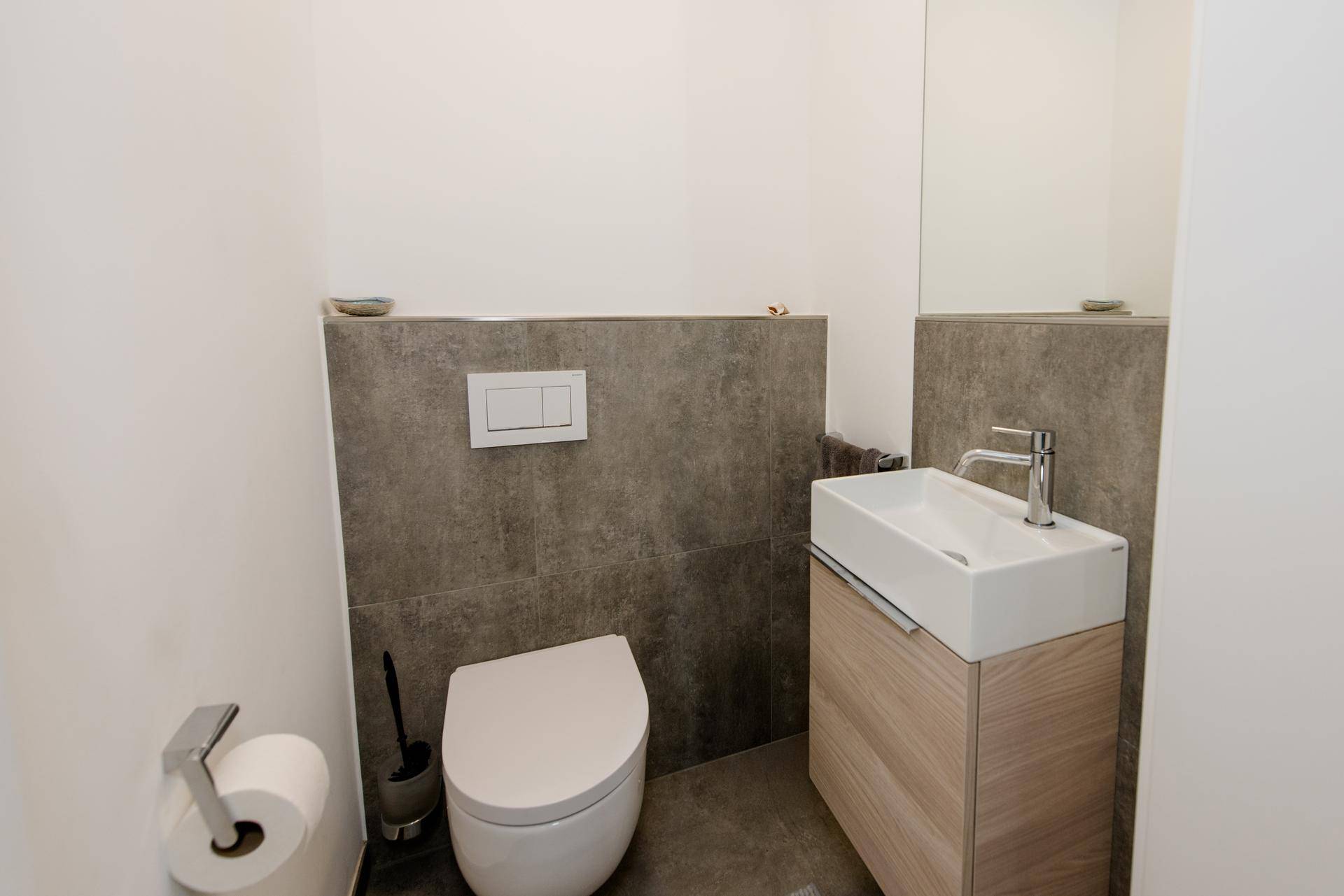 Modernes Gäste-WC mit wandhängender Toilette, kleinem Waschbecken und grauen Fliesen.