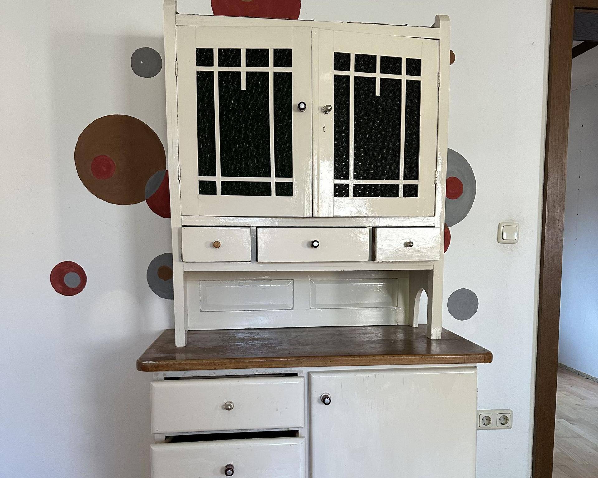 Antiker Holzschrank mit verglasten Türen und Schubladen, dekoriert mit runden Mustern an der Wand.