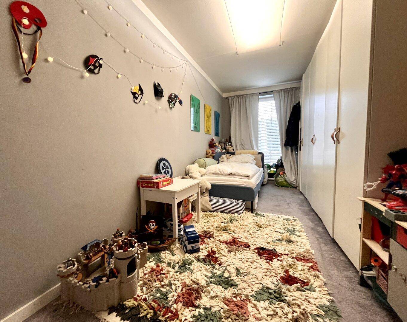 Kinderzimmer mit Einzelbett, weißen Einbauschränken und einem farbenfrohen Teppich.