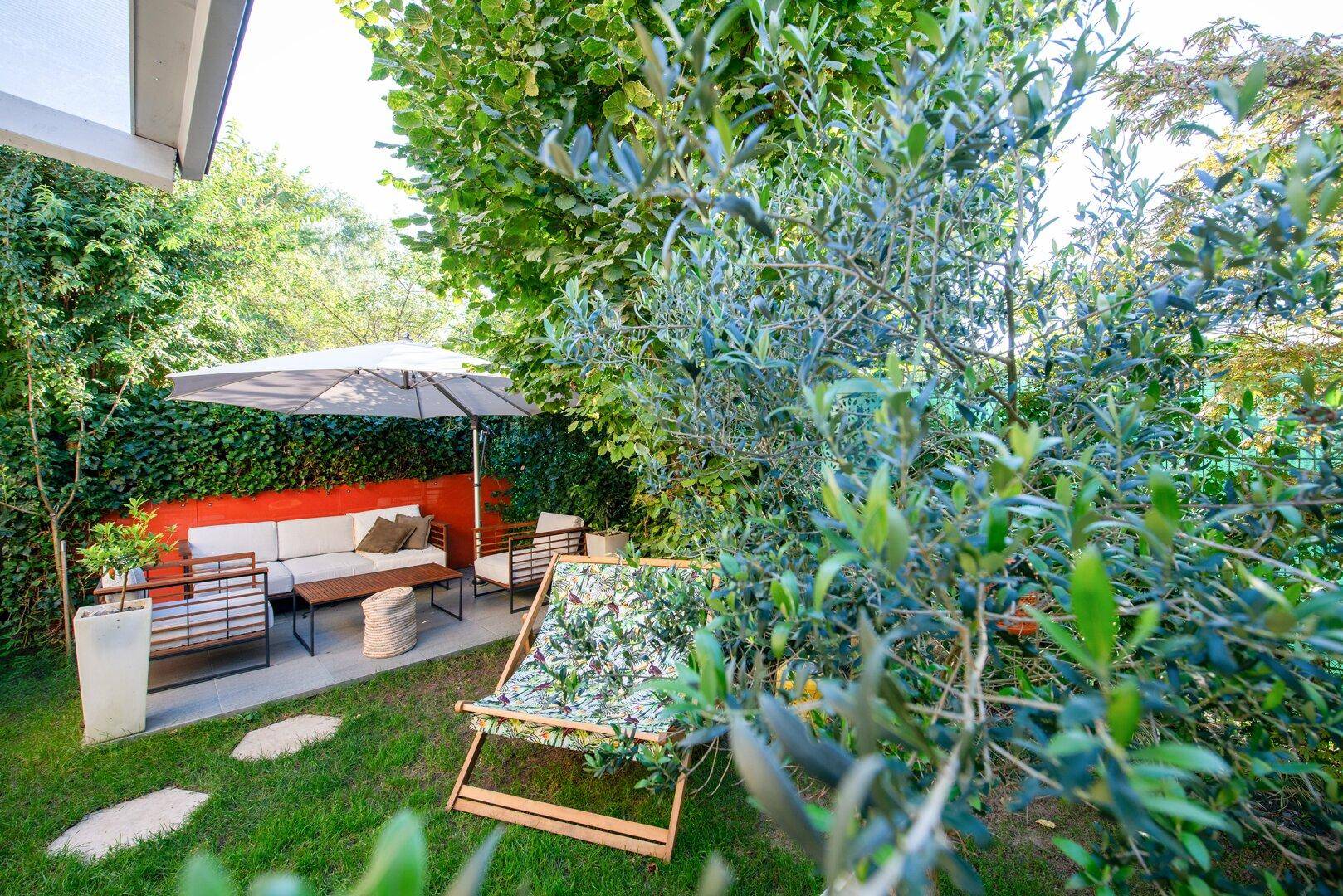 Entspannter Gartenbereich mit Lounge-Möbeln unter einem Sonnenschirm, umgeben von viel Grün.