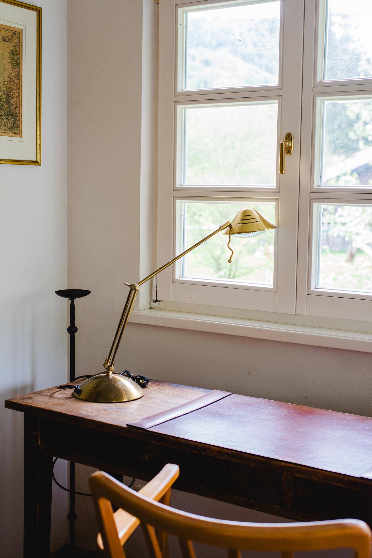 Detailaufnahme eines Schreibtisches mit goldener Lampe vor einem Fenster mit grüner Aussicht.