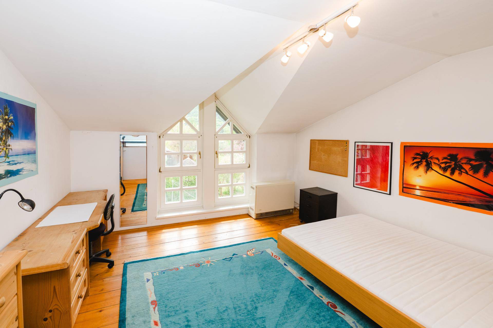 Helles Schlafzimmer mit Holzboden, Schreibtisch und farbenfroher Dekoration unter Dachschrägen.