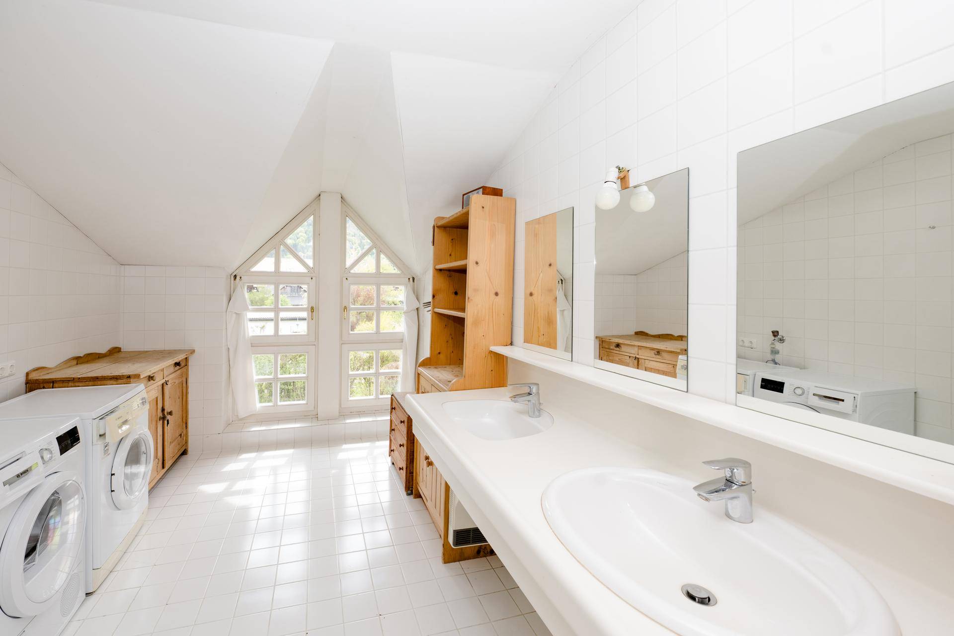 Geräumiges Badezimmer mit Doppelwaschbecken, Waschmaschinen und großen Fenstern unter Dachschrägen.