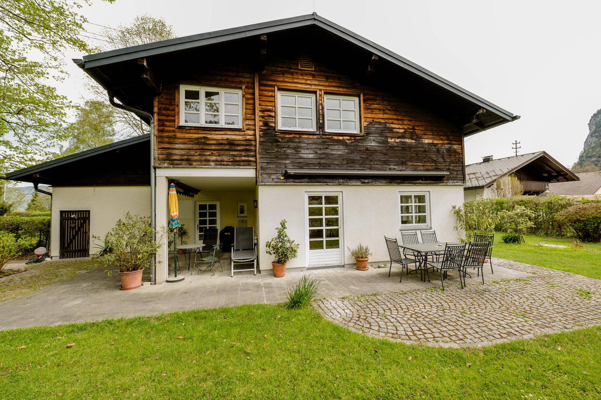 Rückseite des Hauses mit gepflasterter Terrasse, Gartenmöbeln und Blick auf die grüne Umgebung.