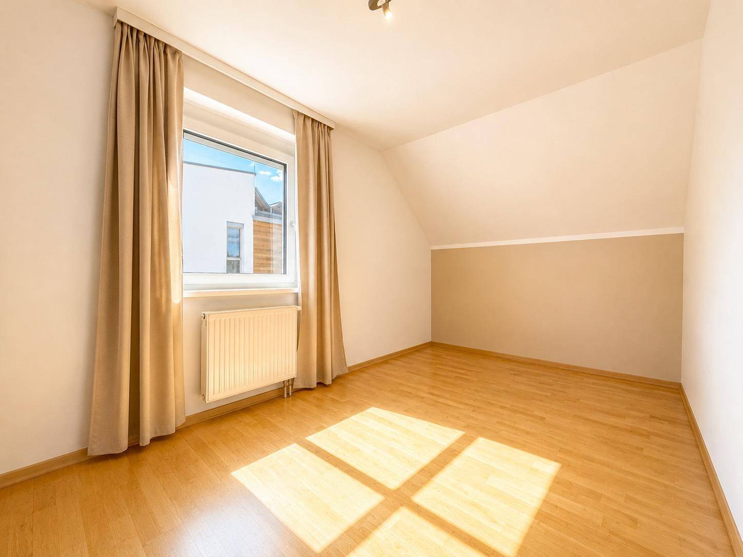 Heller Raum mit Holzboden, großem Fenster und neutralen Wänden, ideal als Schlaf- oder Arbeitszimmer.