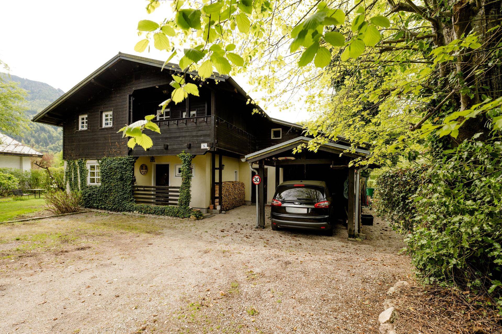 Vorderansicht des Landhauses mit Carport und Zufahrt, umgeben von Bäumen und Sträuchern.