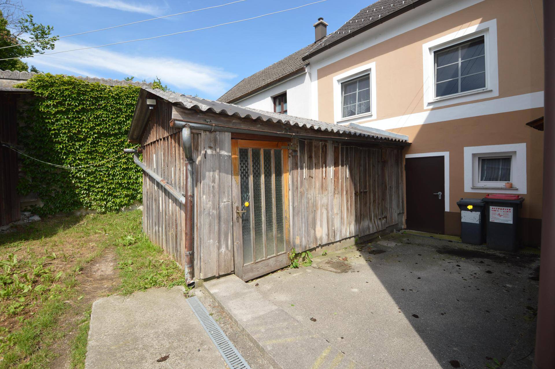 Hinterhofbereich mit einem einfachen Holzschuppen und einer begrünten Wand, umgeben von Gras.