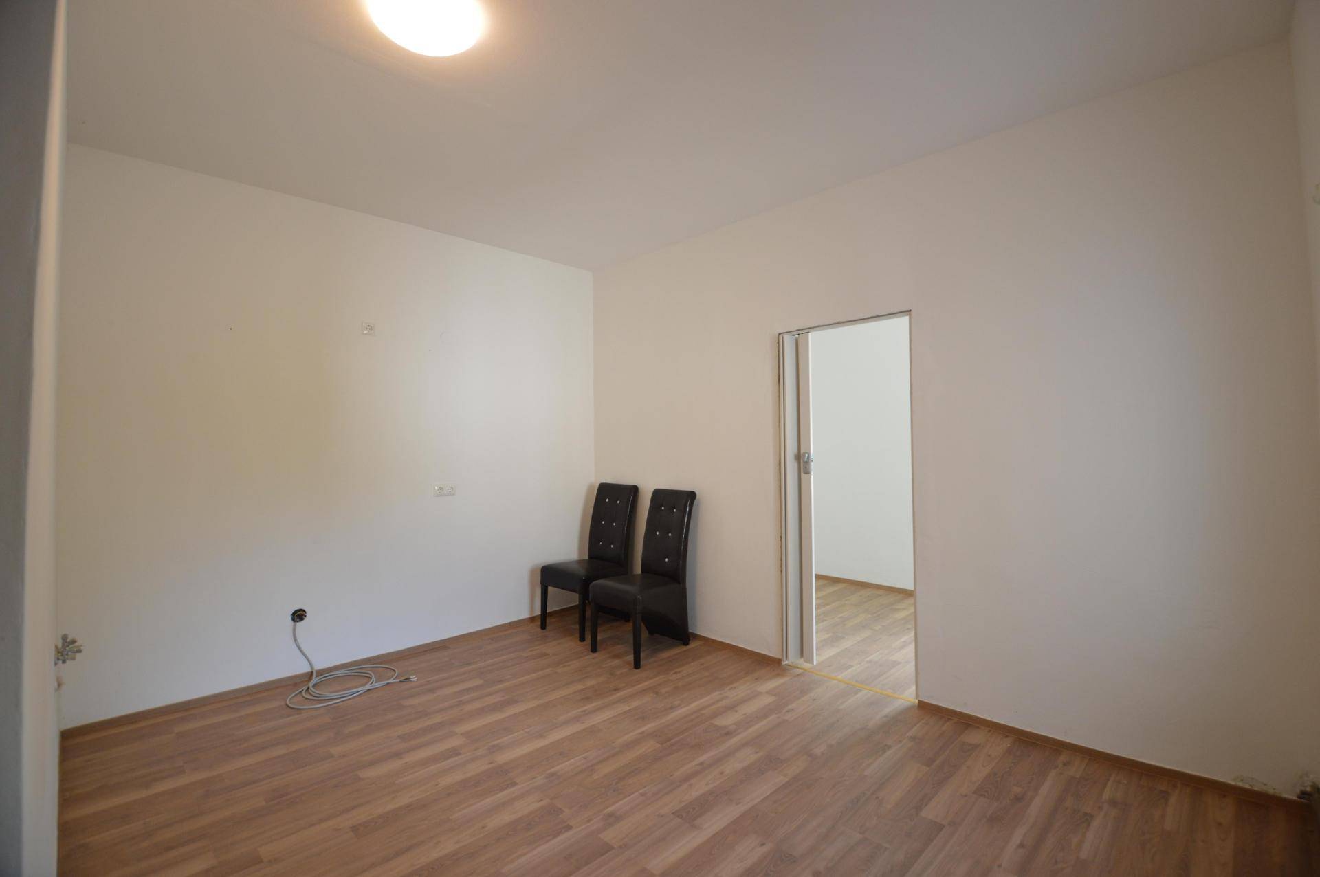 Heller, leerer Raum mit Laminatboden und einer Tür, die zu einem weiteren Zimmer führt.