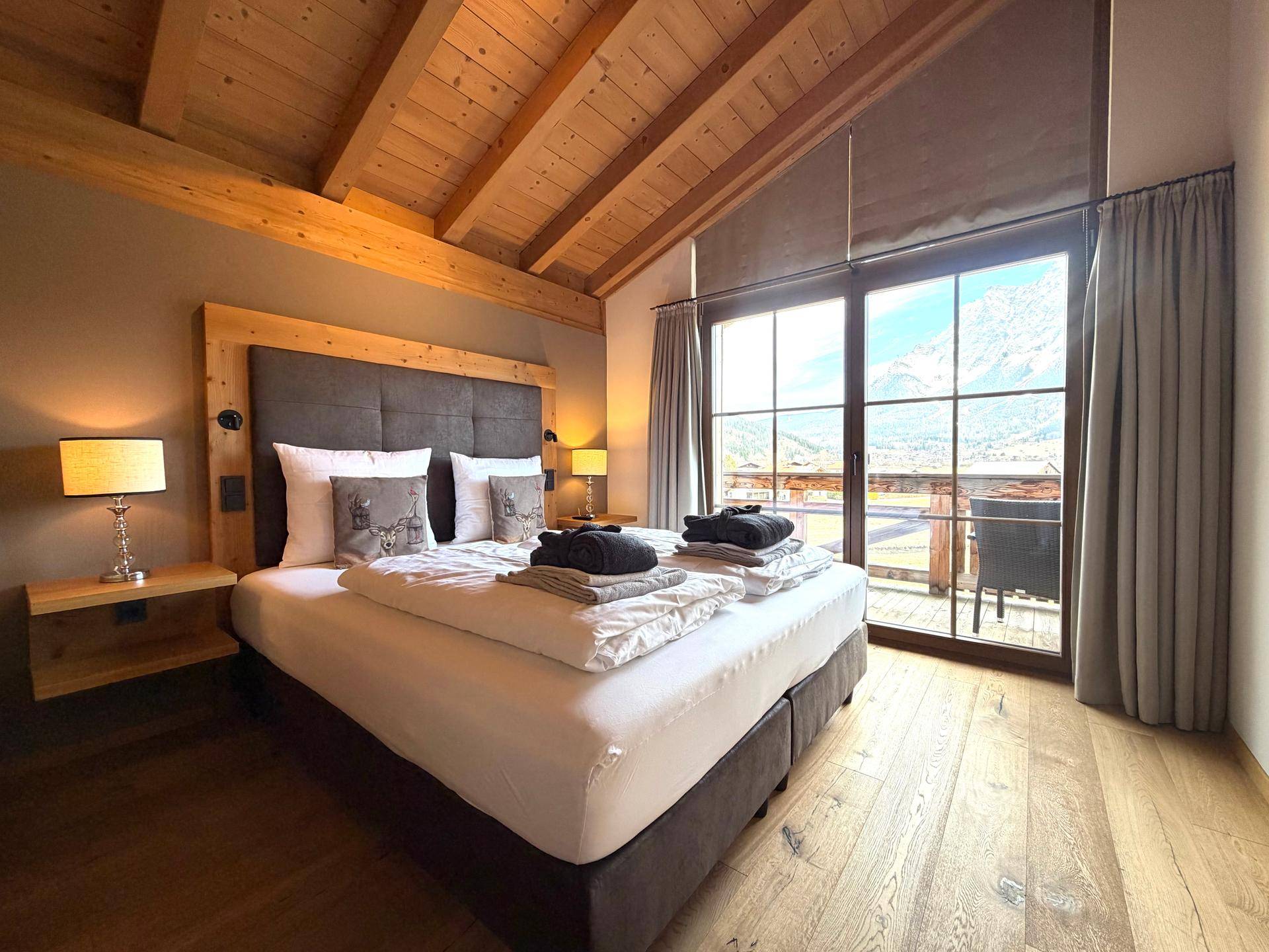 Helles Schlafzimmer mit Holzbalkendecke, Doppelbett und Zugang zu einem Balkon mit Bergblick.