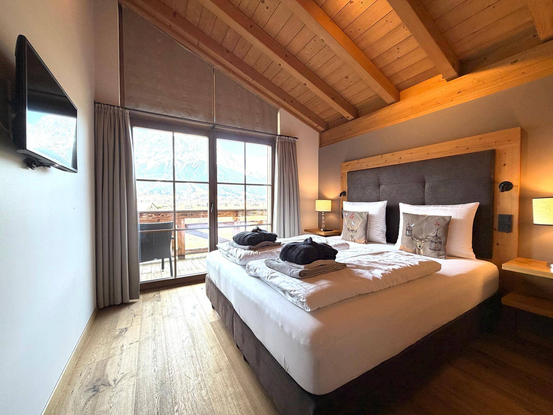 Modernes Schlafzimmer mit Holzbalkendecke, Doppelbett, Flachbildfernseher und Bergblick.