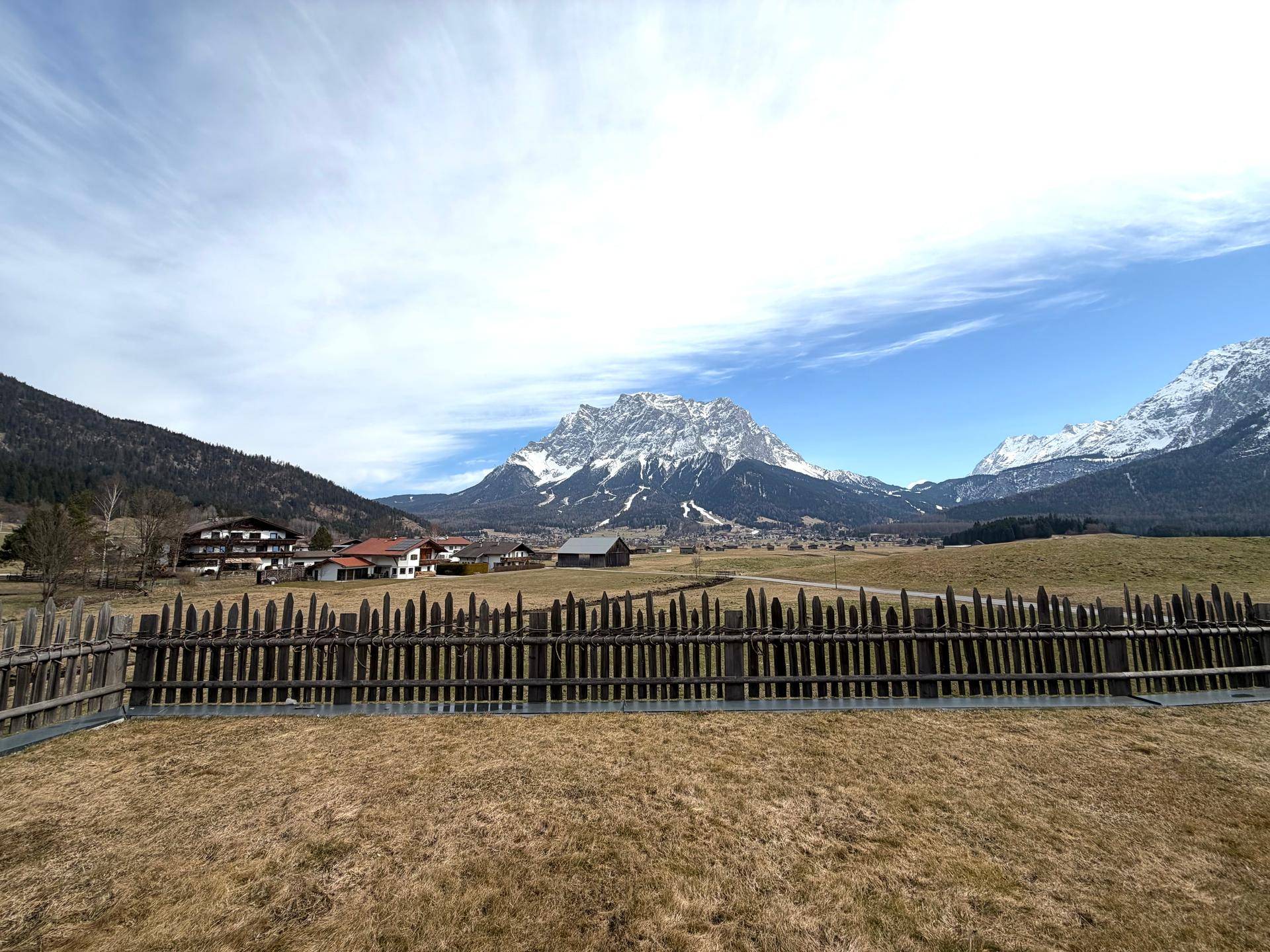 Weitläufiger Blick auf schneebedeckte Berge und grüne Felder unter blauem Himmel.