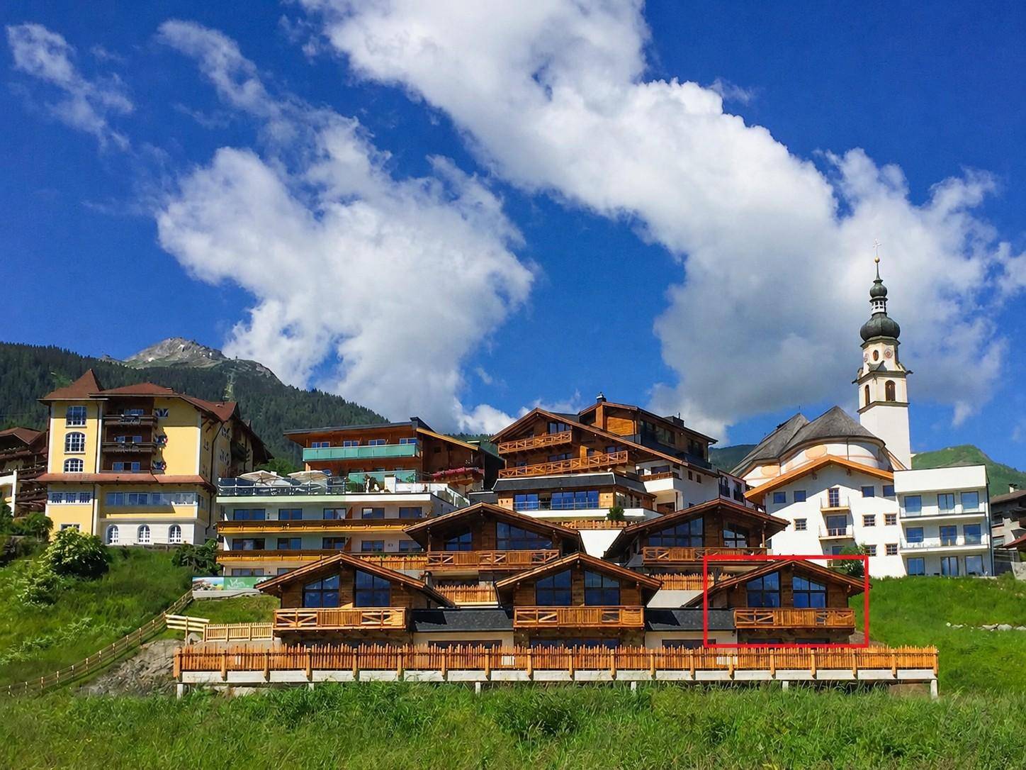 Gruppe von modernen Holzchalets in einer malerischen Berglandschaft mit Kirchturm im Hintergrund.