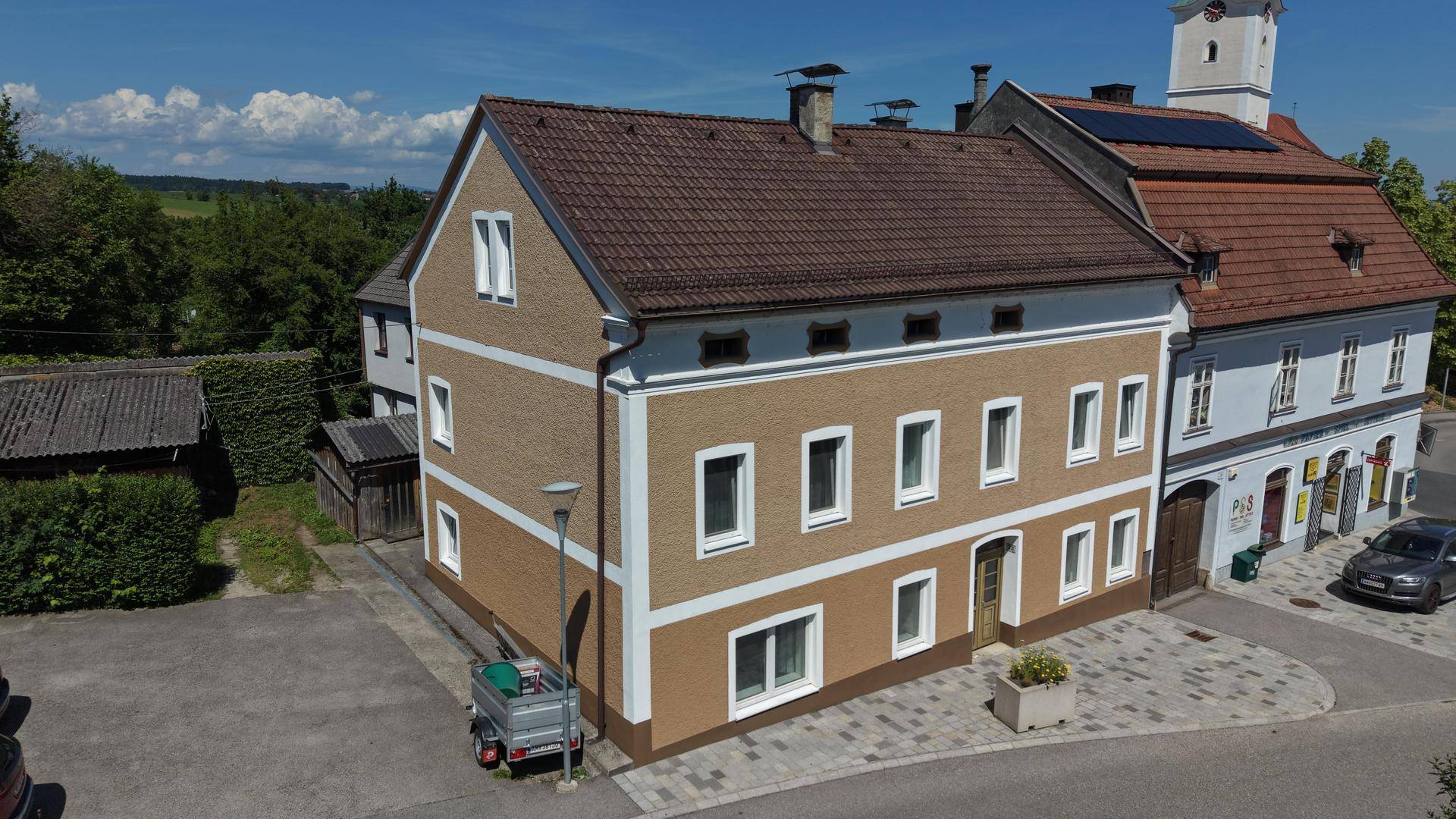 Luftaufnahme des Gebäudes mit ockerfarbener Fassade und braunem Ziegeldach, umgeben von weiteren Häusern.