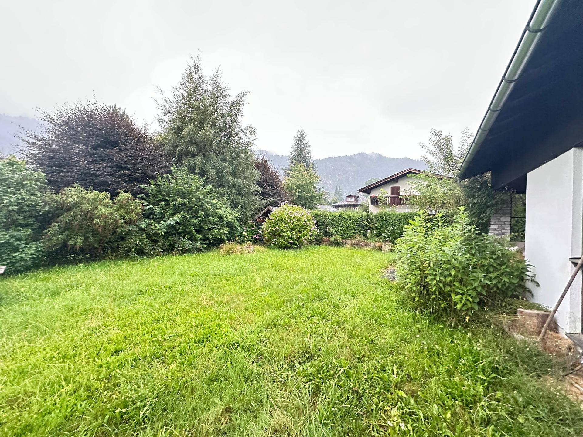 Grüner Garten mit Rasenfläche, Sträuchern und Bäumen vor einer bergigen Kulisse.