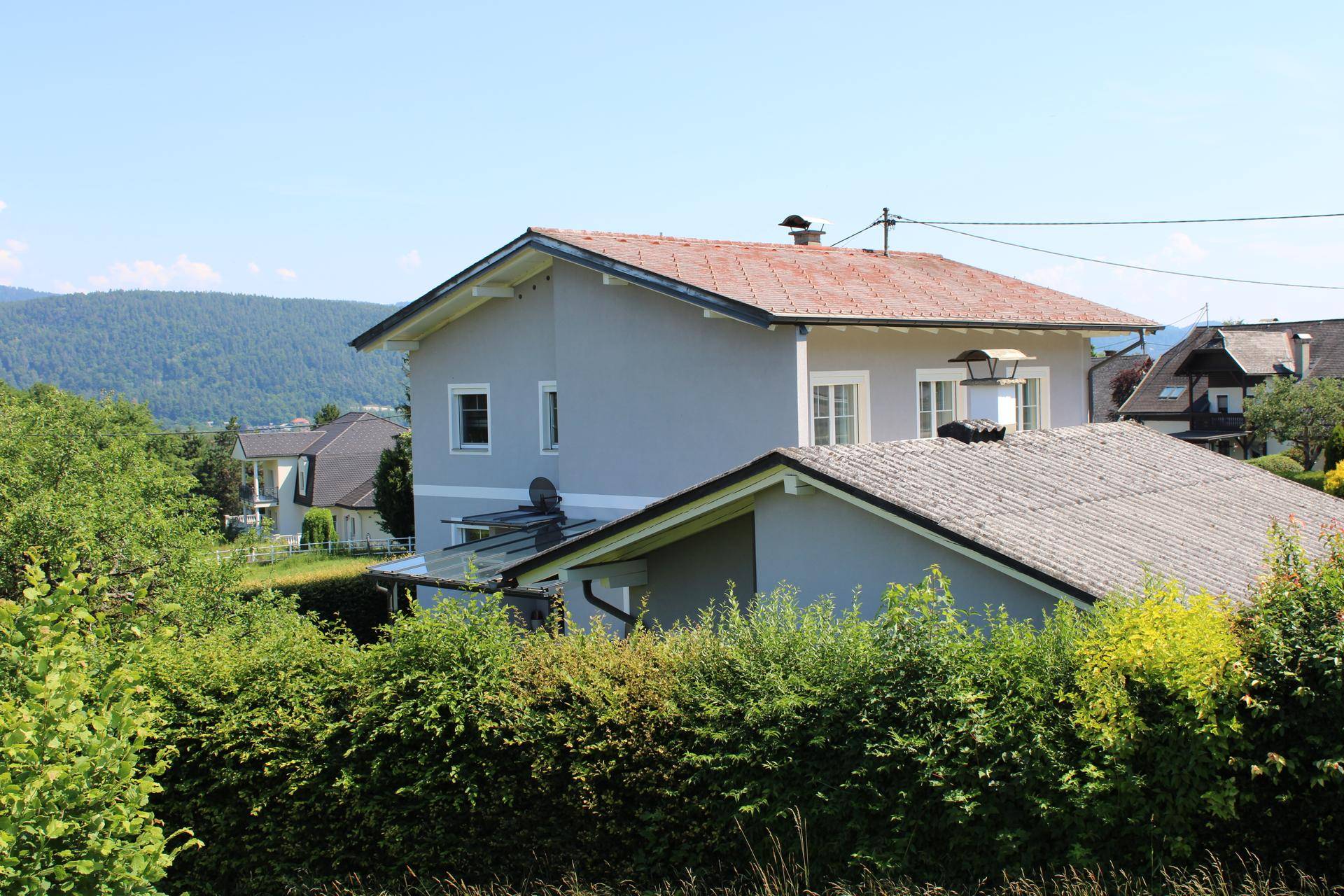 Seitenansicht eines grauen Hauses mit Satteldach, umgeben von Bäumen und Hecken.