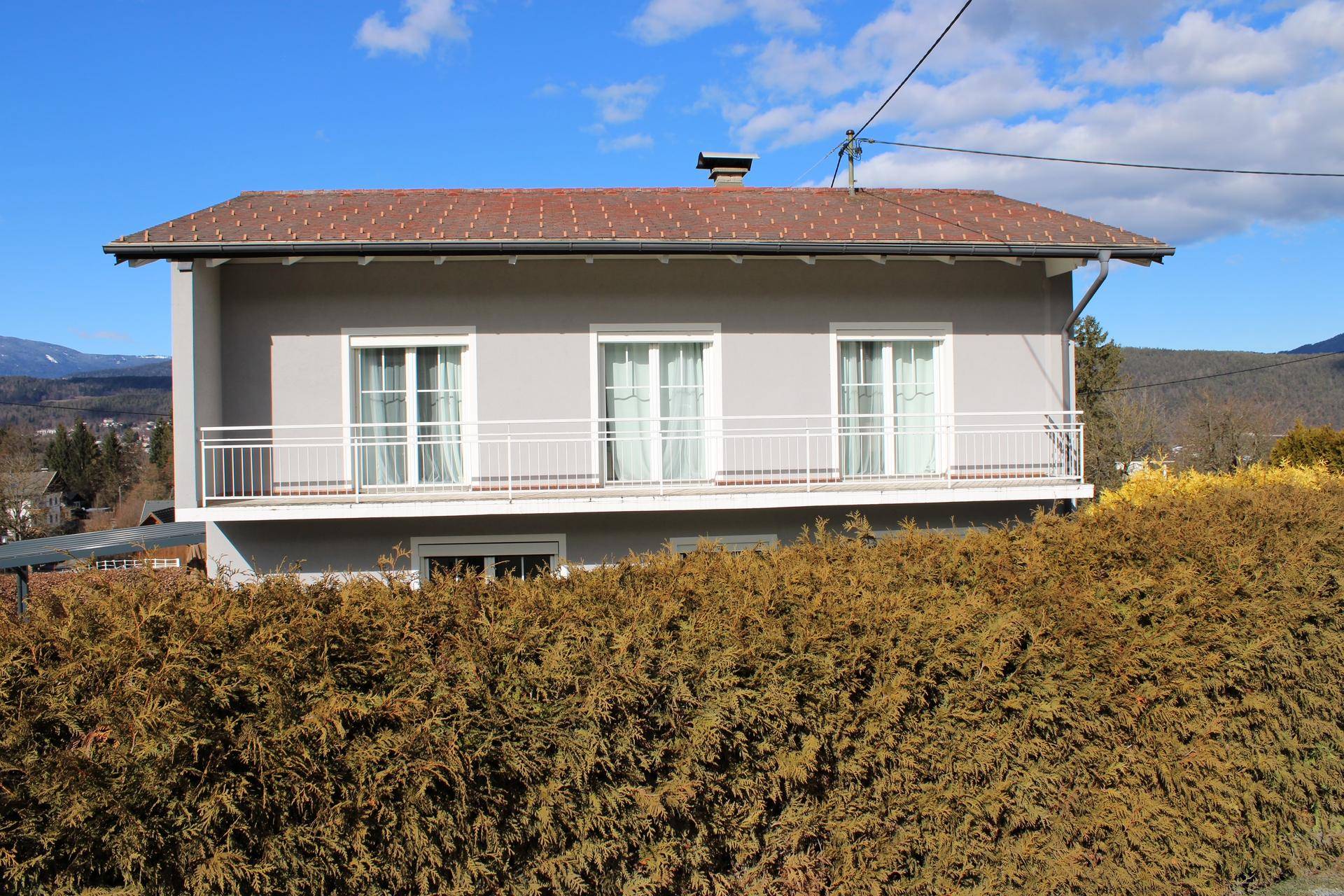 Vorderansicht eines grauen Hauses mit Balkon, teilweise von einer Hecke verdeckt.