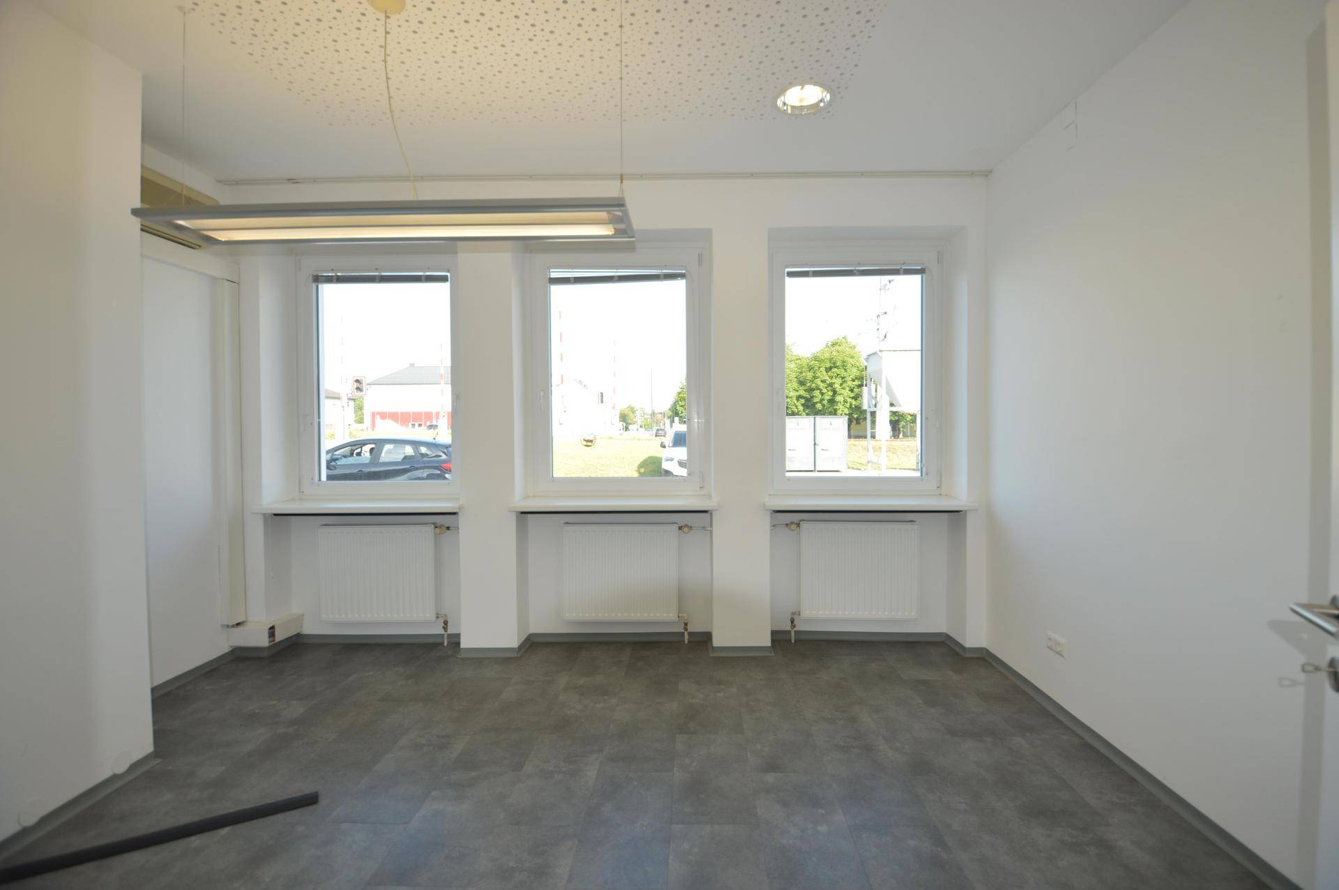 Geräumiges Büro mit drei Fenstern, die viel Tageslicht hereinlassen, und Heizkörpern darunter.