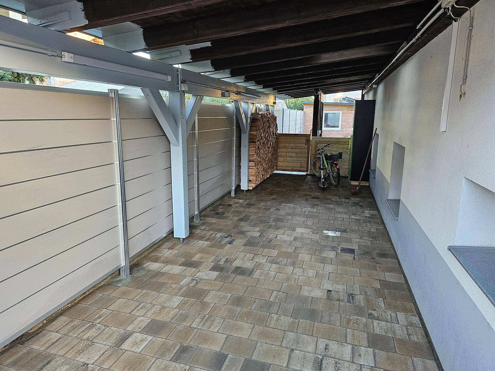 Überdachter Stellplatz mit gepflastertem Boden und Holzstapel im Hintergrund.