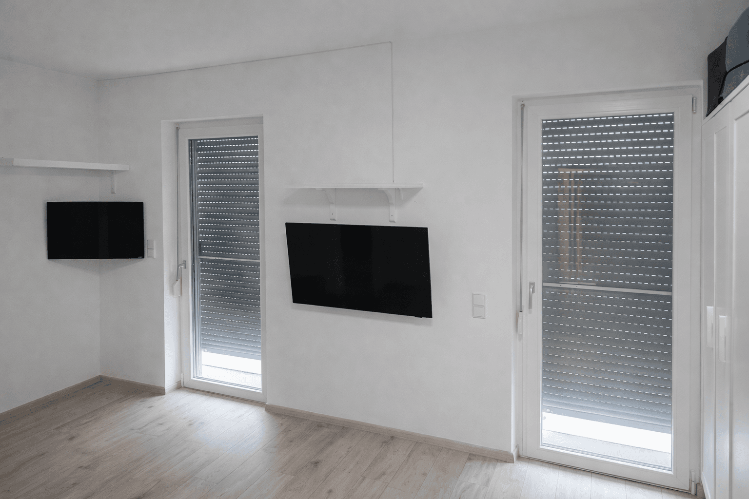 Heller Raum mit zwei Fenstern und Rollläden sowie Wandfernsehern.