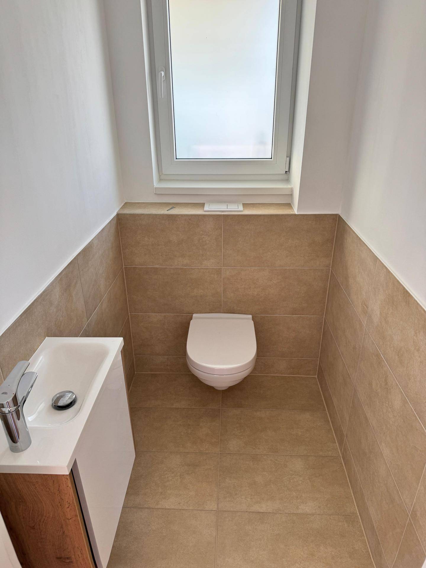 Modernes Gäste-WC mit beigefarbenen Fliesen, wandhängendem WC und kleinem Waschbecken mit Fenster.