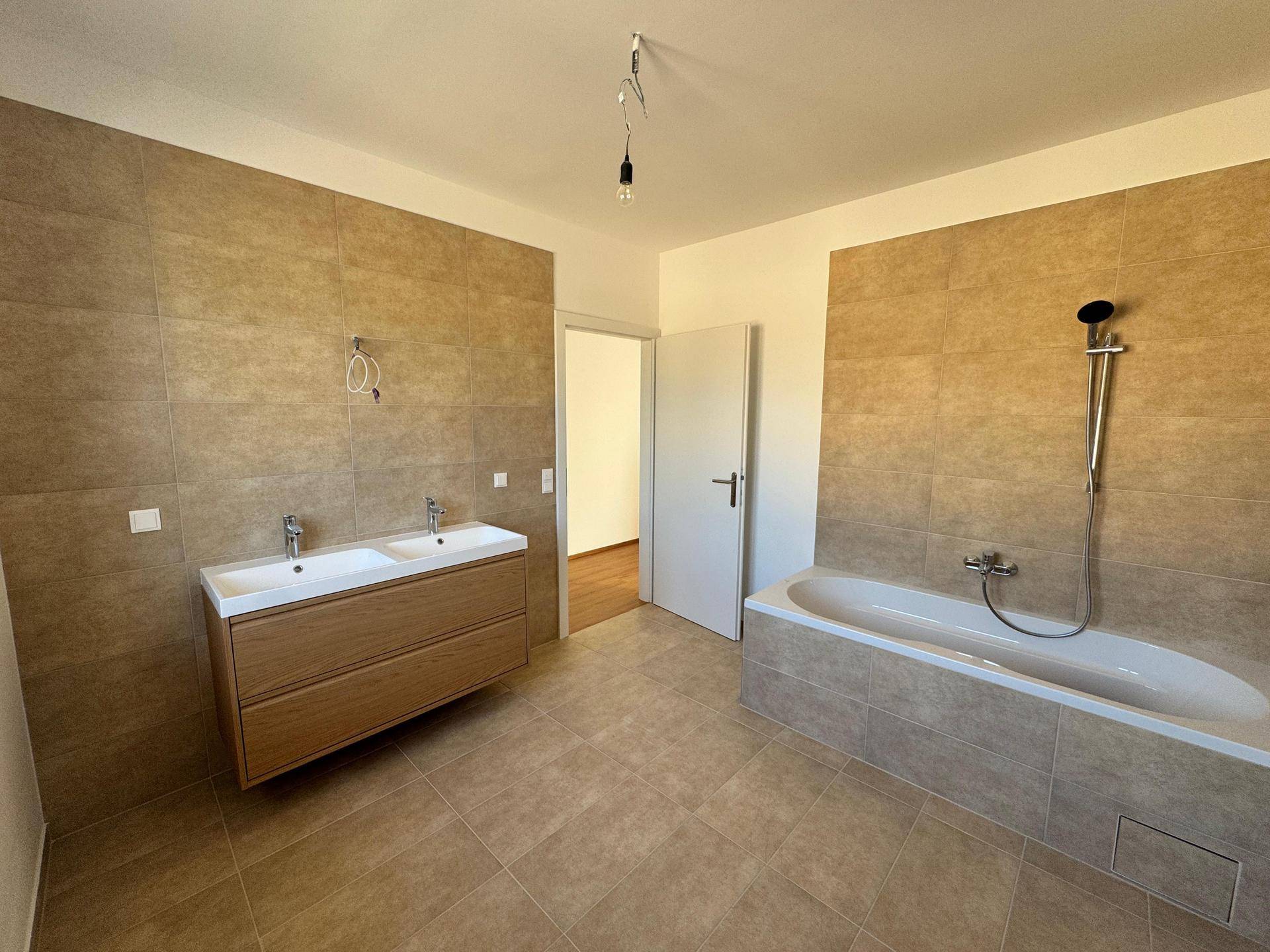 Geräumiges Badezimmer mit beigefarbenen Fliesen, Doppelwaschbecken und Badewanne mit Duscharmatur.