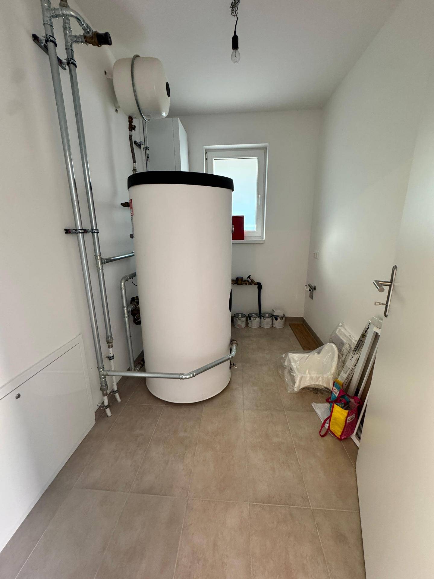 Hauswirtschaftsraum mit Warmwasserspeicher, Heizungsrohren und kleinem Fenster.
