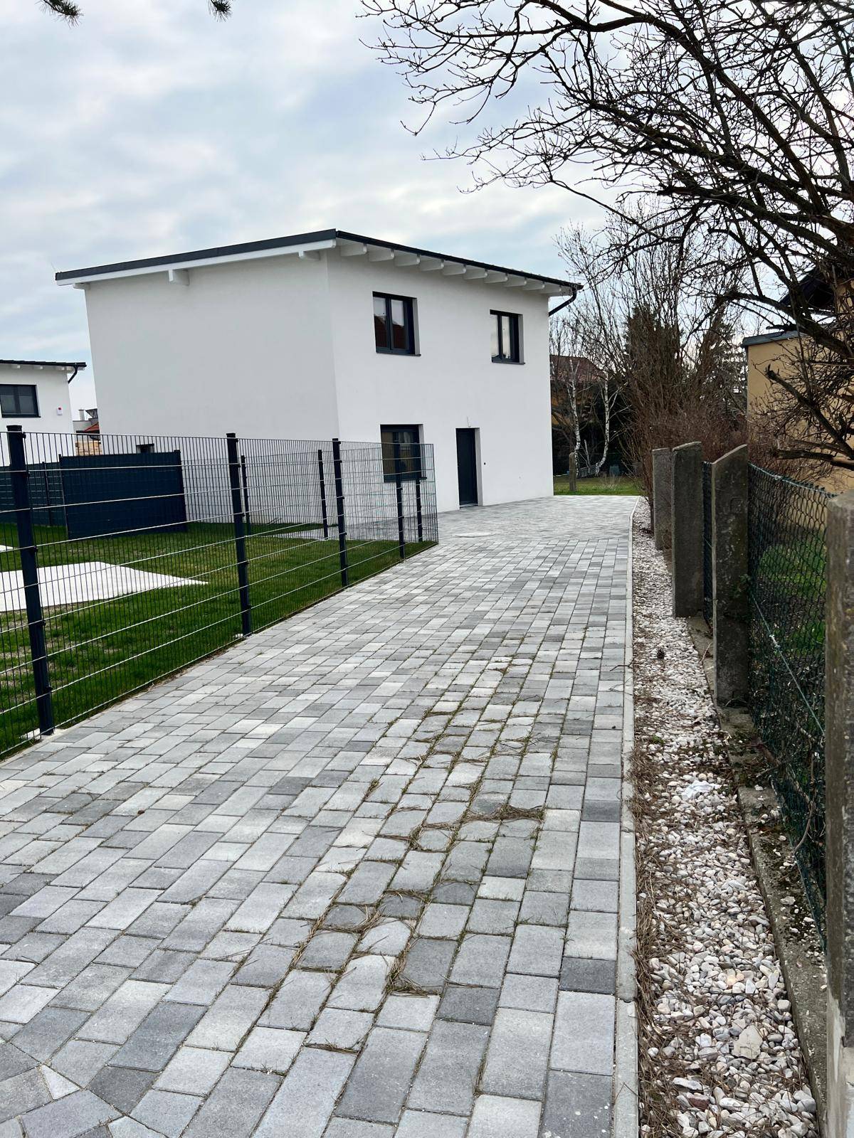 Modernes Einfamilienhaus mit gepflasterter Einfahrt und umzäuntem Gartenbereich an einem bewölkten Tag.
