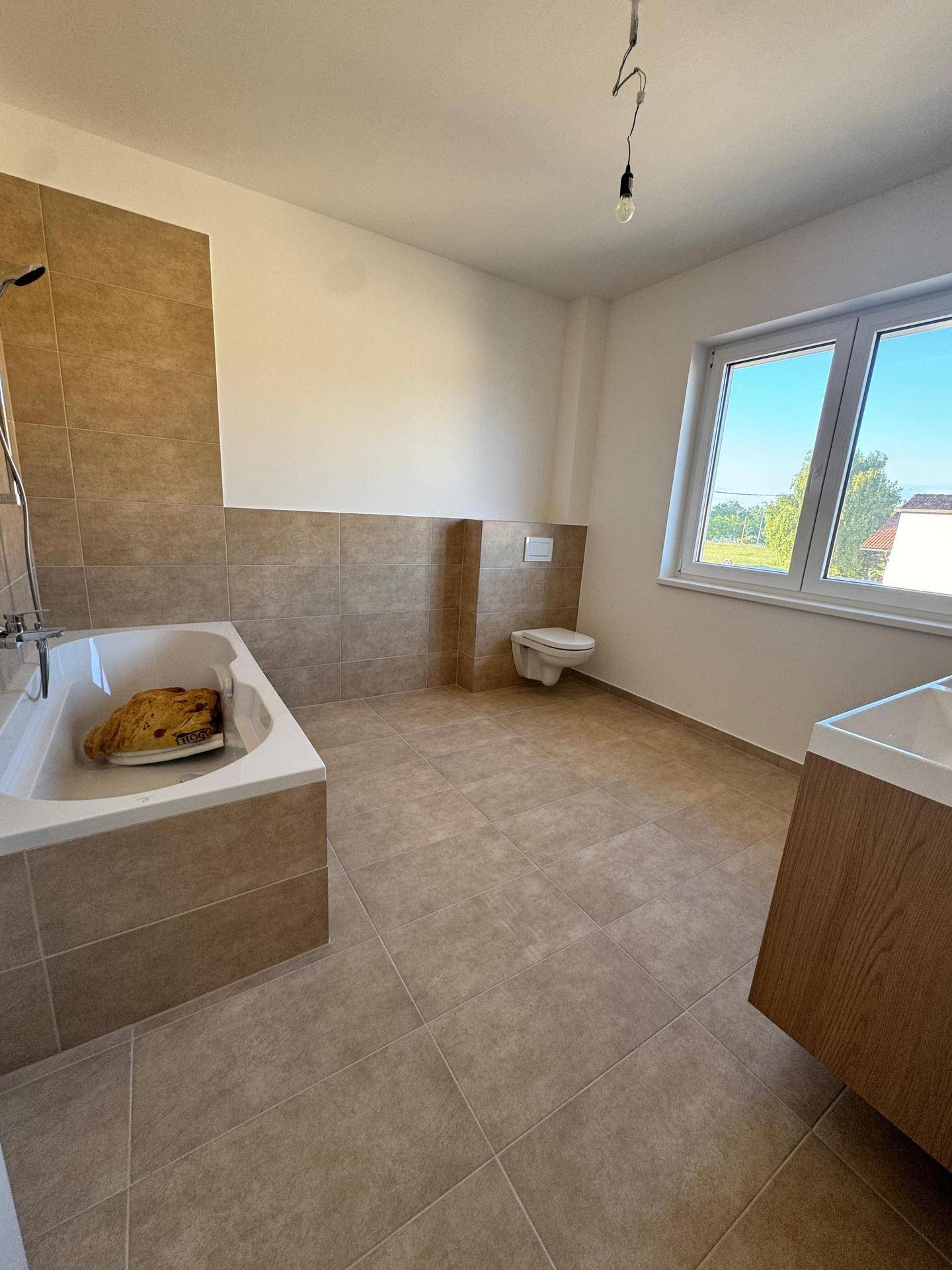 Helles Badezimmer mit beigefarbenen Fliesen, Badewanne, wandhängendem WC und Fenster mit Ausblick.