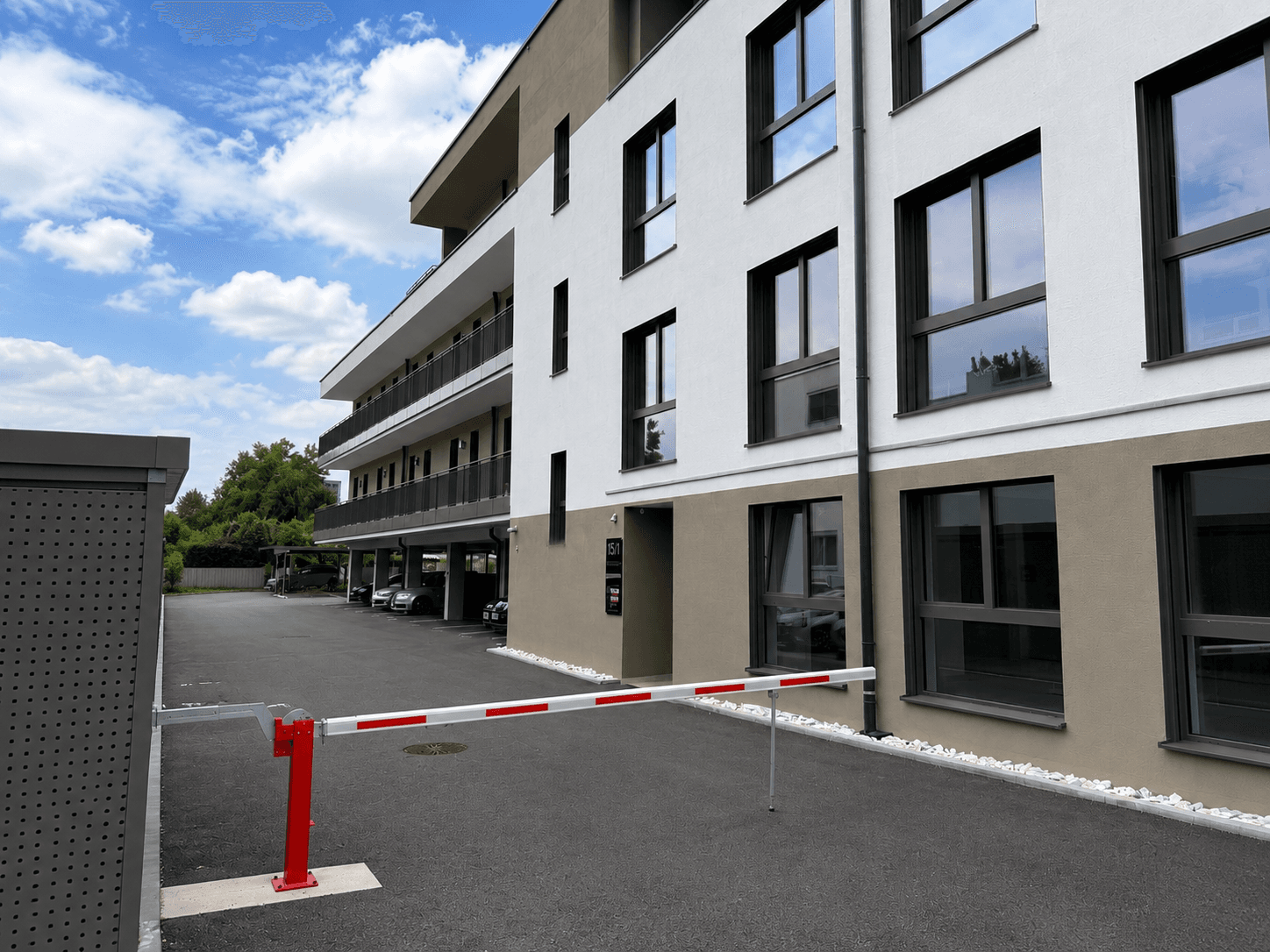 Seitenansicht des modernen Gebäudes mit Parkplatz, Schranke, Balkonen und Rollläden unter blauem Himmel.