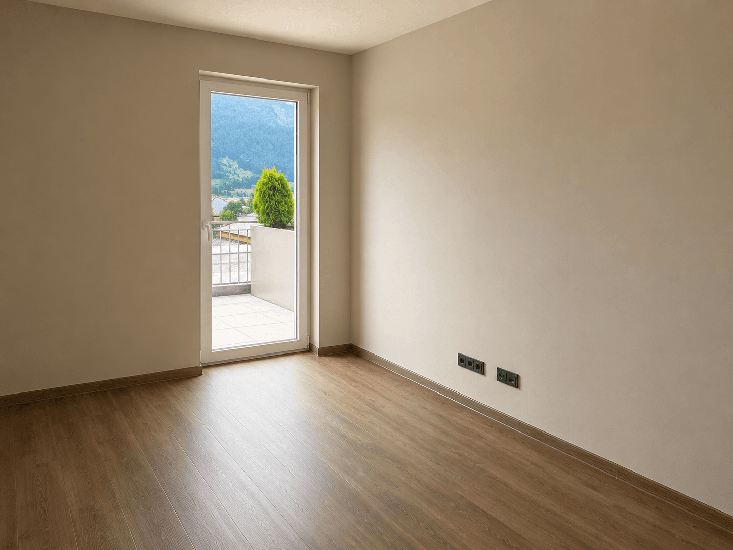 Leeres Zimmer mit Laminatboden und Balkonzugang, bietet einen Blick ins Grüne.