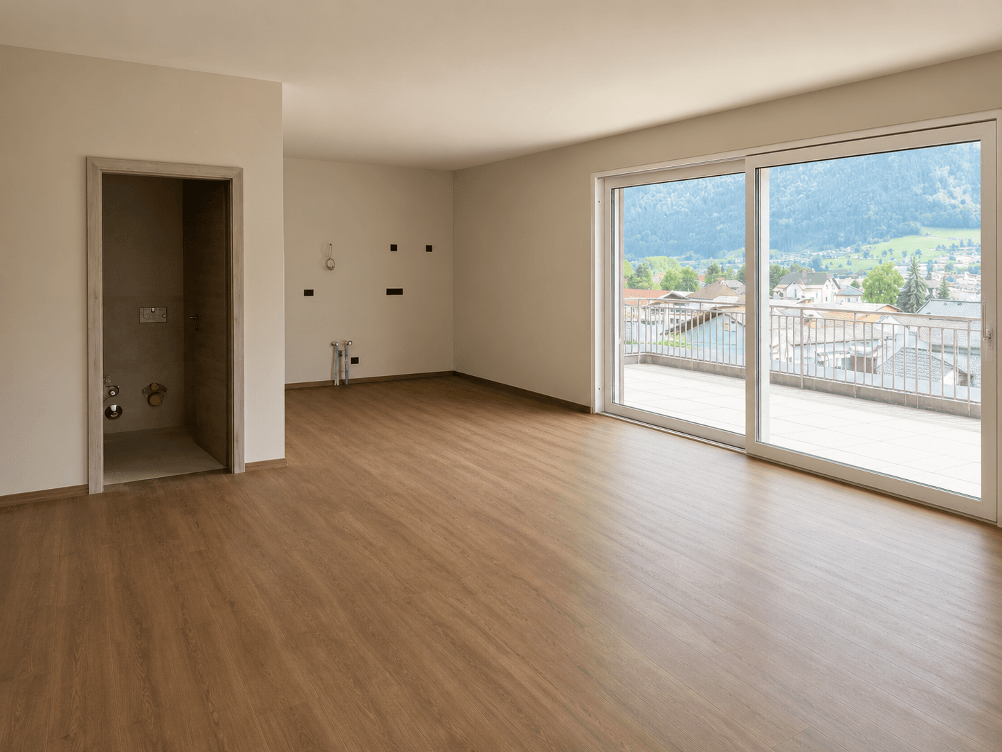 Heller, leerer Wohnraum mit Laminatboden, großen Fenstern und Zugang zur Terrasse mit Bergblick.