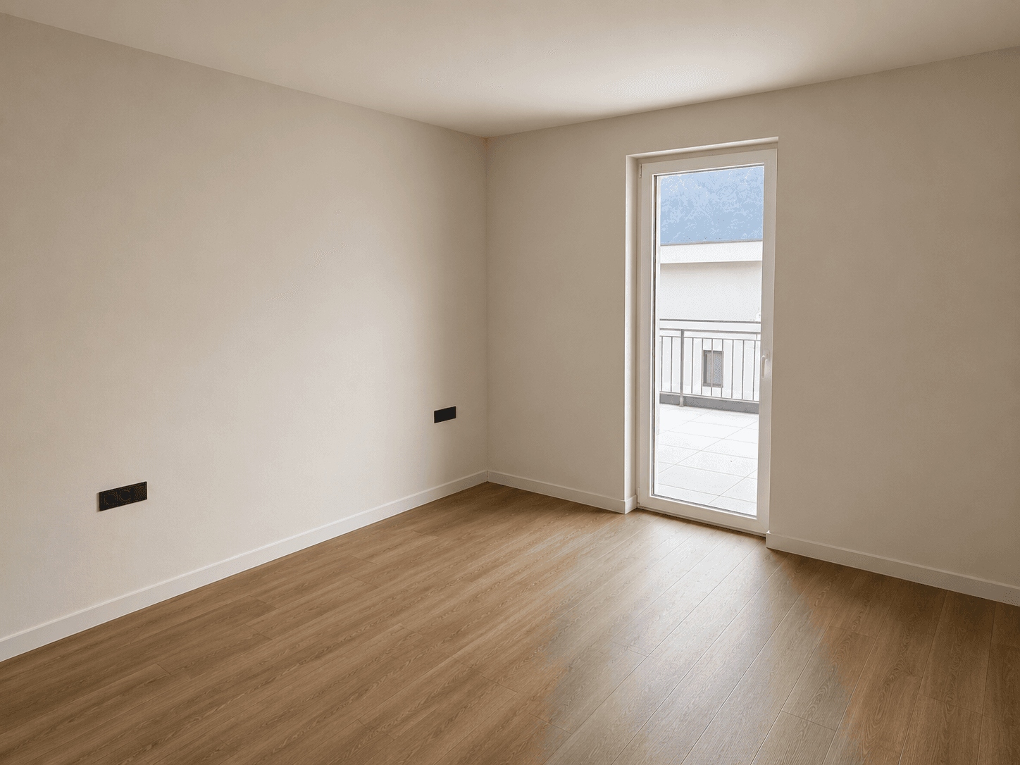 Unmöbliertes Zimmer mit Laminatboden und einer Tür, die auf einen kleinen Balkon führt.