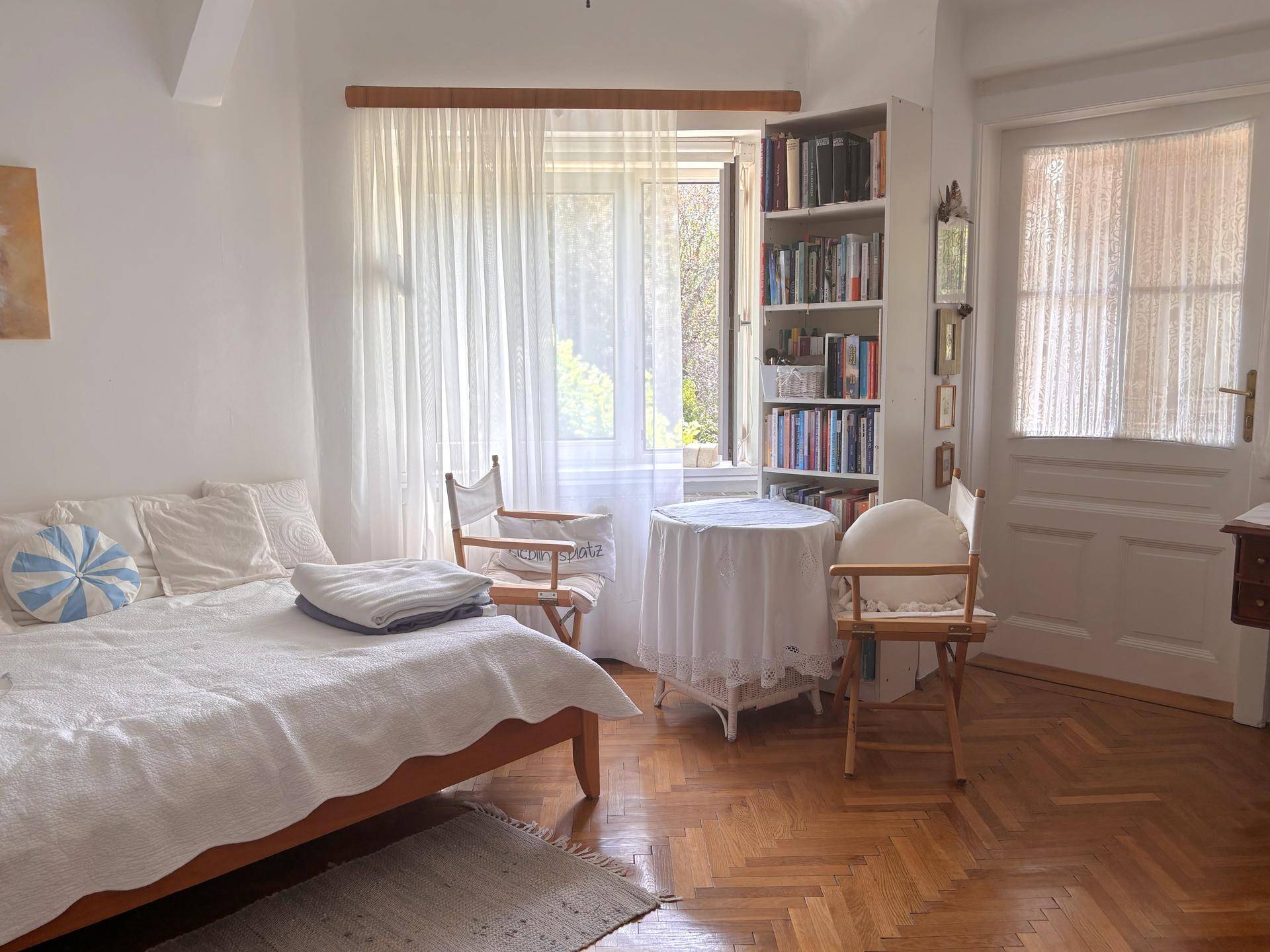 Helles Schlafzimmer mit Parkettboden, Bücherregal und Zugang zu einem kleinen Tischbereich.