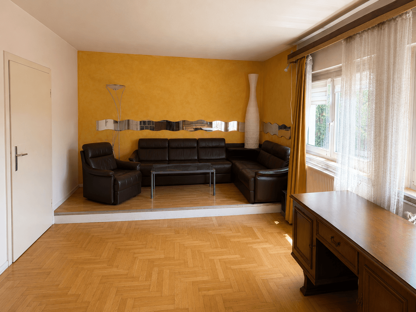 Geräumiges Wohnzimmer mit Parkettboden, gelber Akzentwand und erhöhter Sitzecke.
