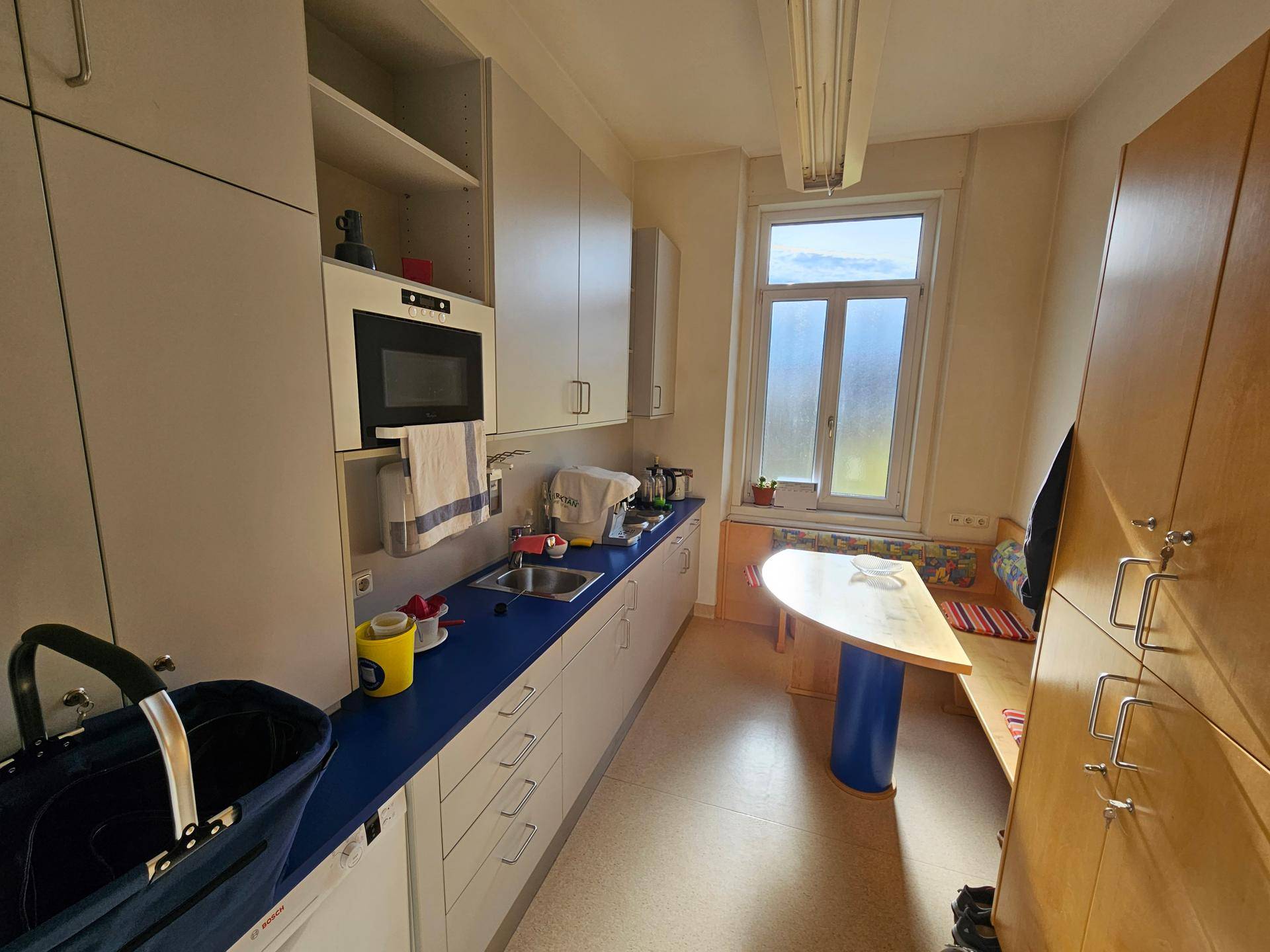 Schmale Küche mit hellgrauen Schränken, blauer Arbeitsplatte und einem kleinen Essbereich am Fenster.