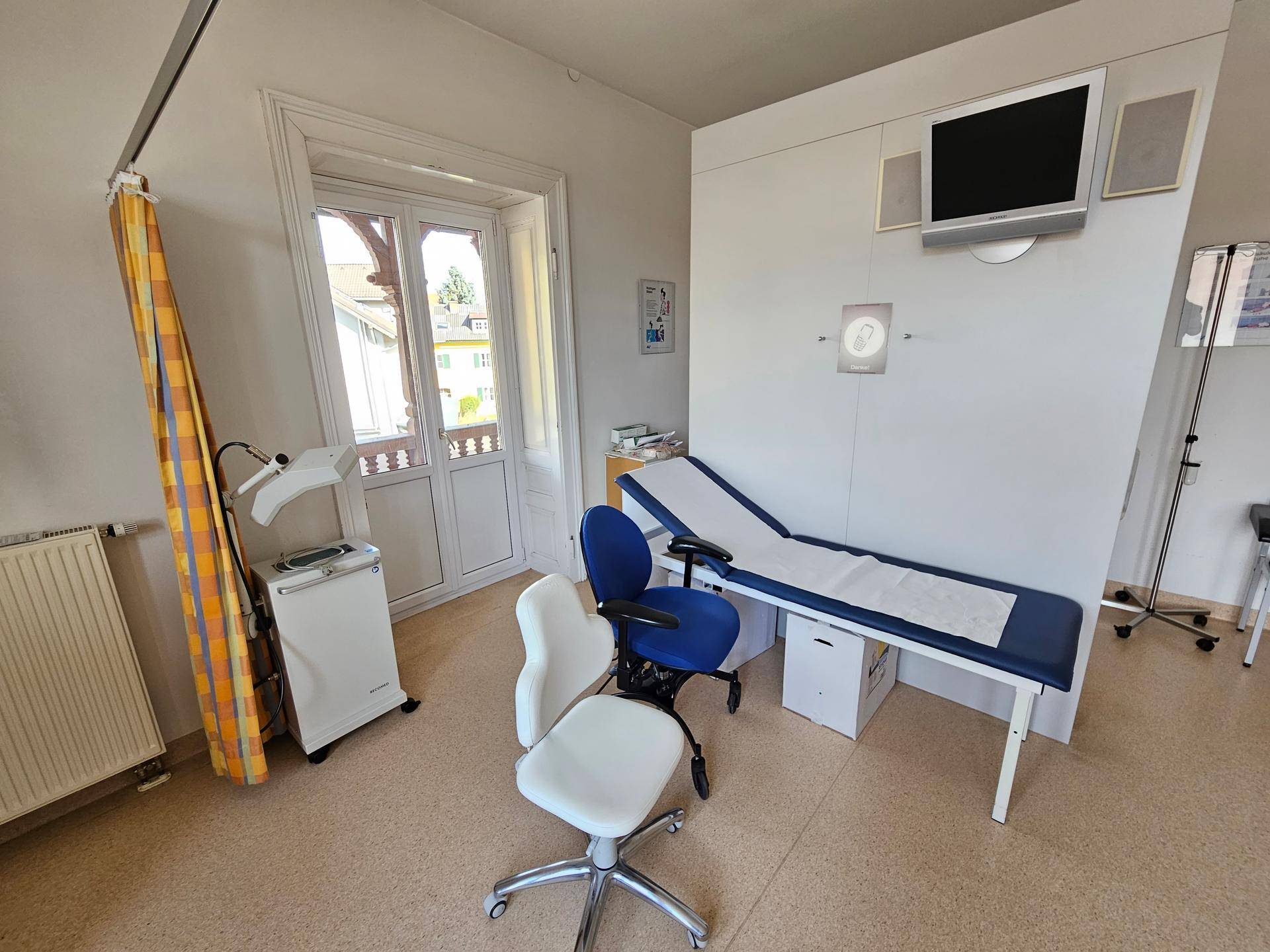 Behandlungszimmer mit medizinischer Ausstattung, einem Fenster und Zugang zu einem kleinen Balkon.