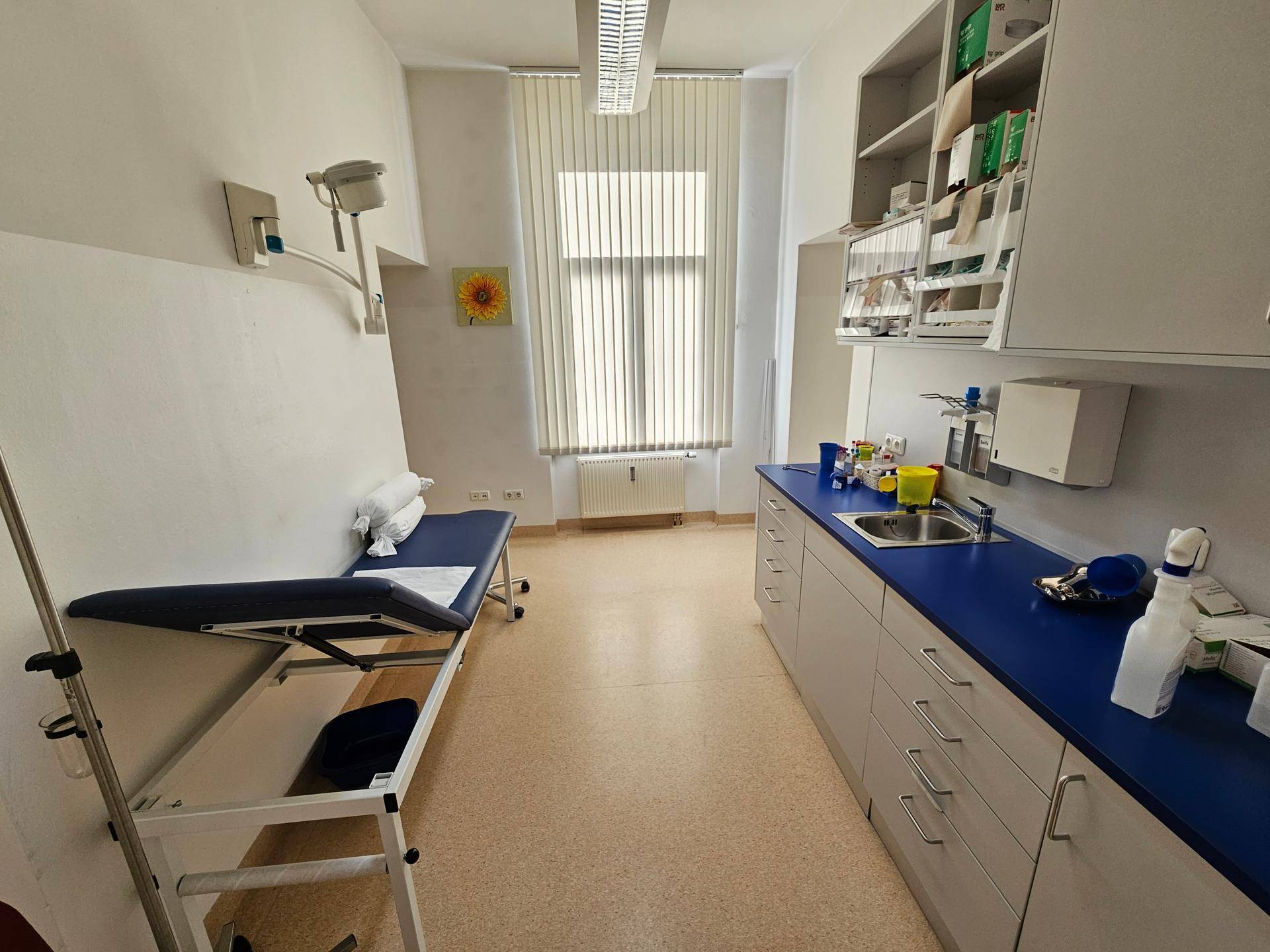 Behandlungsraum mit Untersuchungsliege, Waschbecken und Medizinschränken.