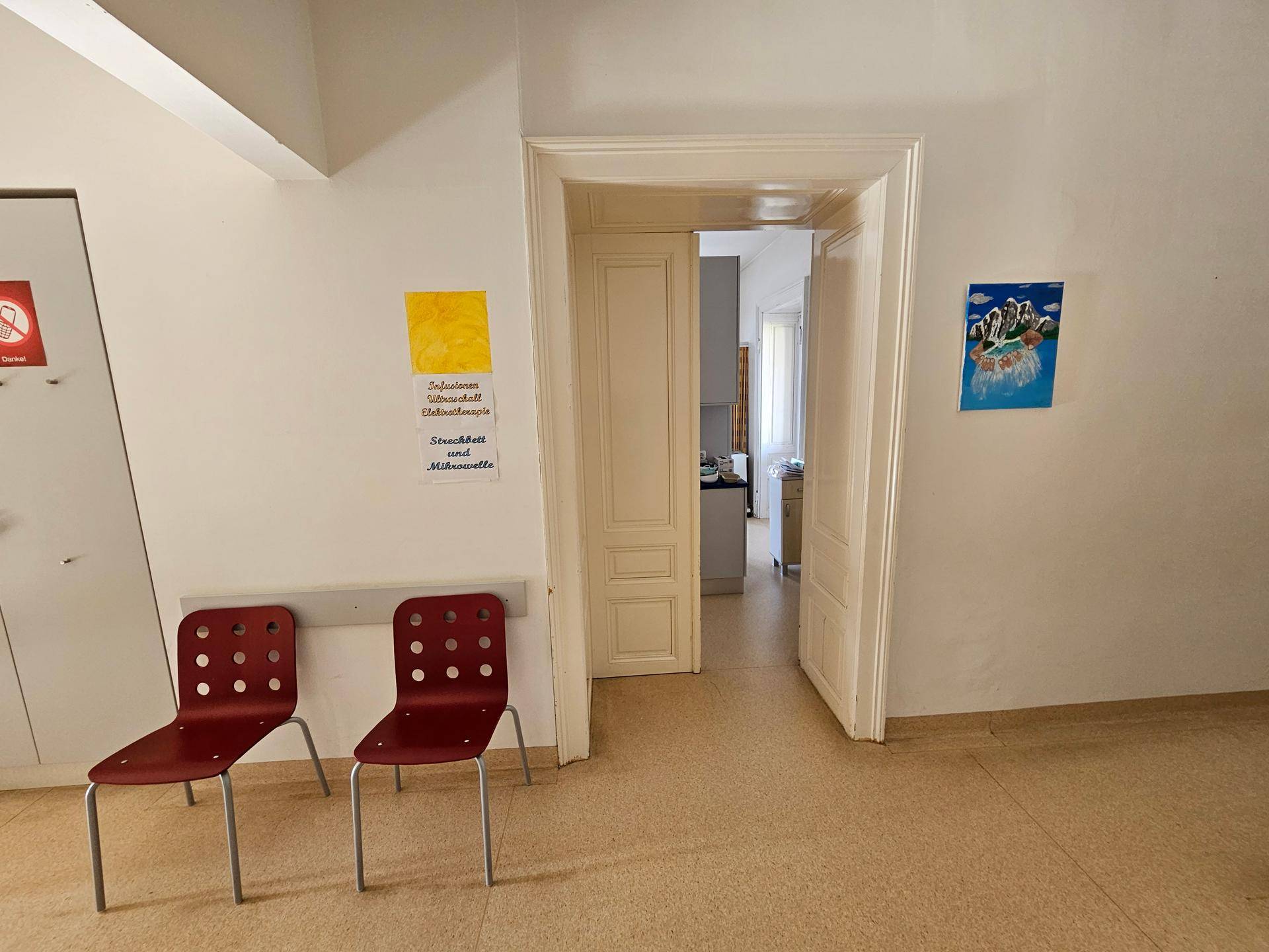 Heller Flur oder Wartebereich mit zwei roten Stühlen und einem offenen Durchgang zu weiteren Räumen.