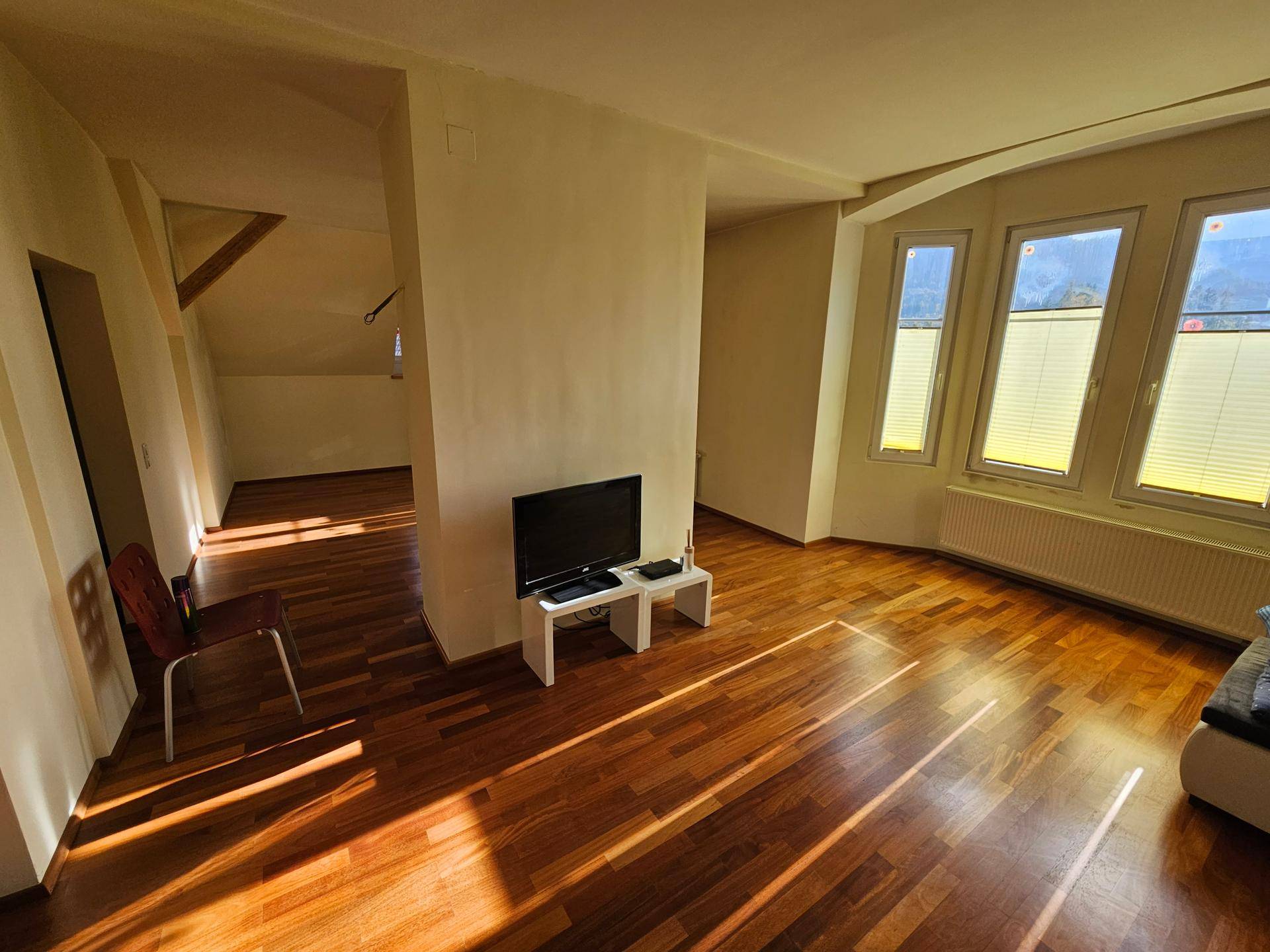 Heller Wohnbereich mit Holzboden, großen Fenstern und sichtbaren Holzbalken im Hintergrund.