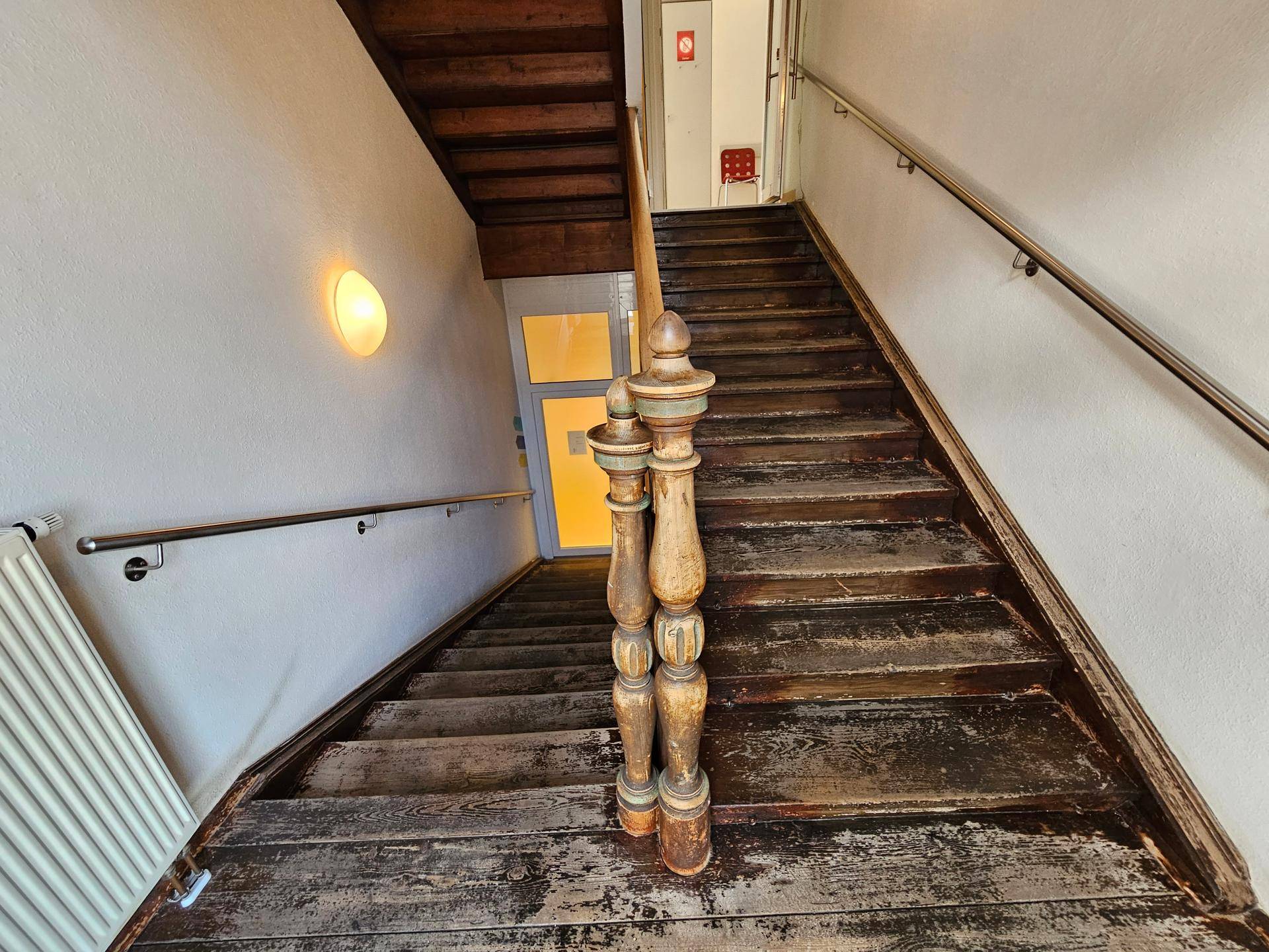 Abgenutzte Holztreppe mit Geländer und Heizkörper, die in die oberen Stockwerke führt.