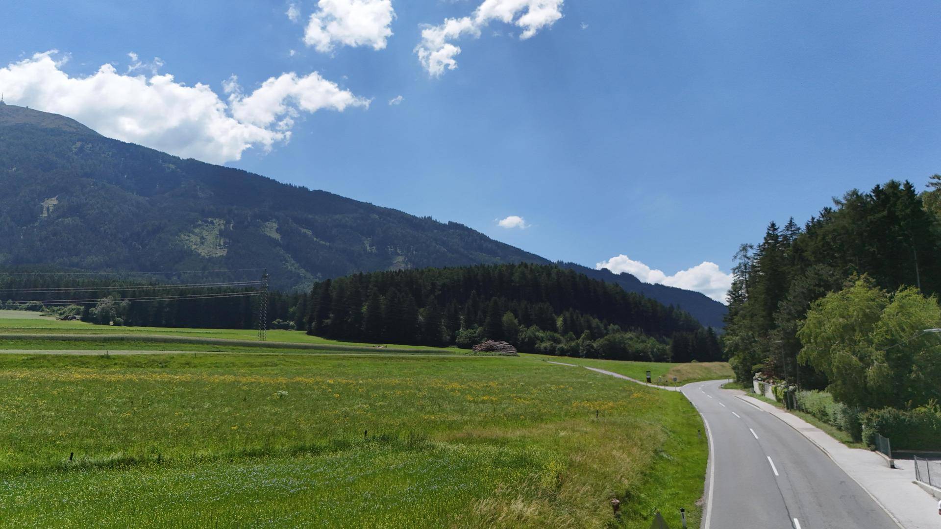 Eine Landstraße schlängelt sich durch grüne Felder, umgeben von Wald und Bergen in der Ferne.