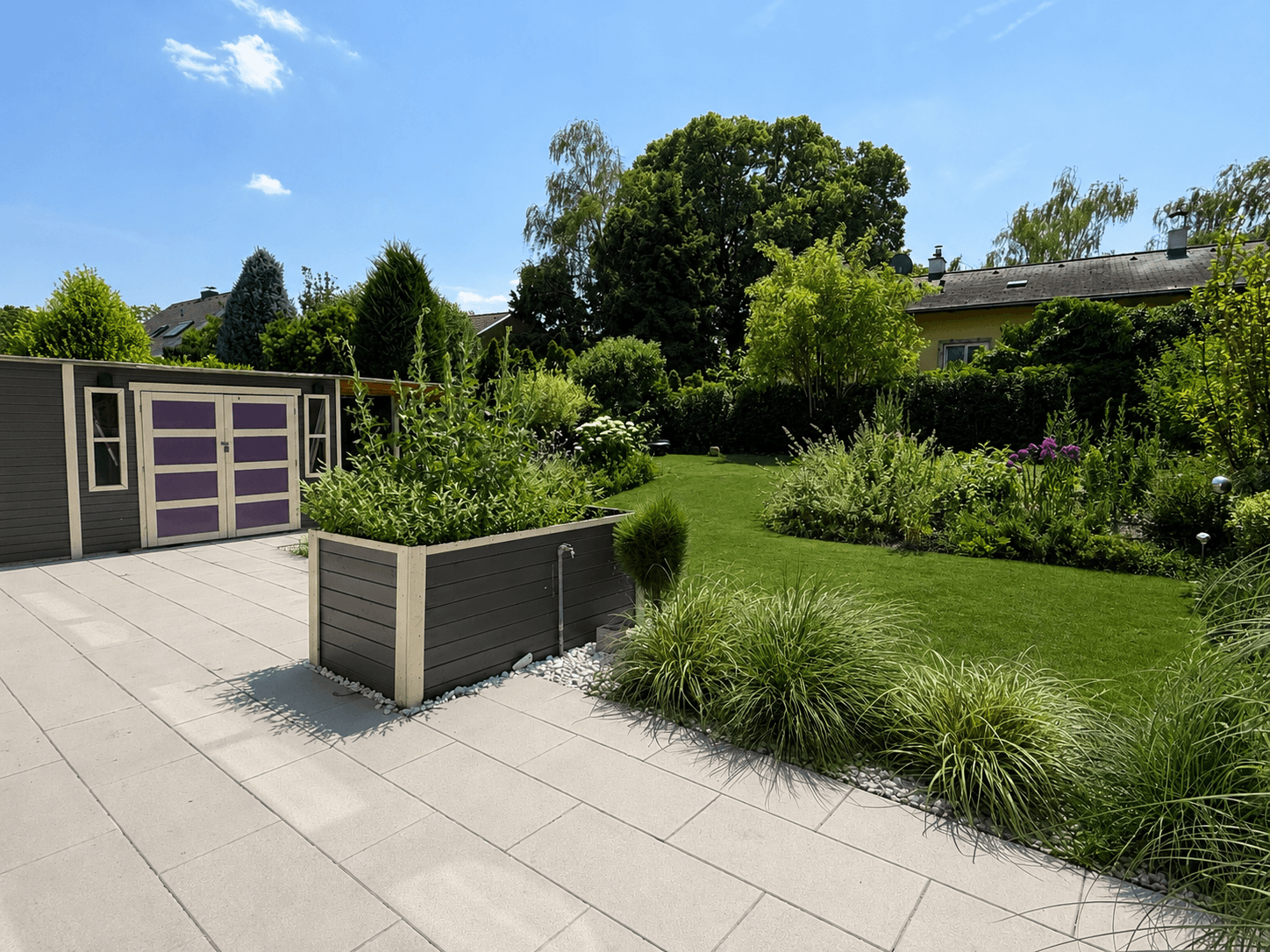 Gepflasterte Terrasse mit Hochbeeten und einem Gartenhaus, angrenzend an einen grünen Rasen.