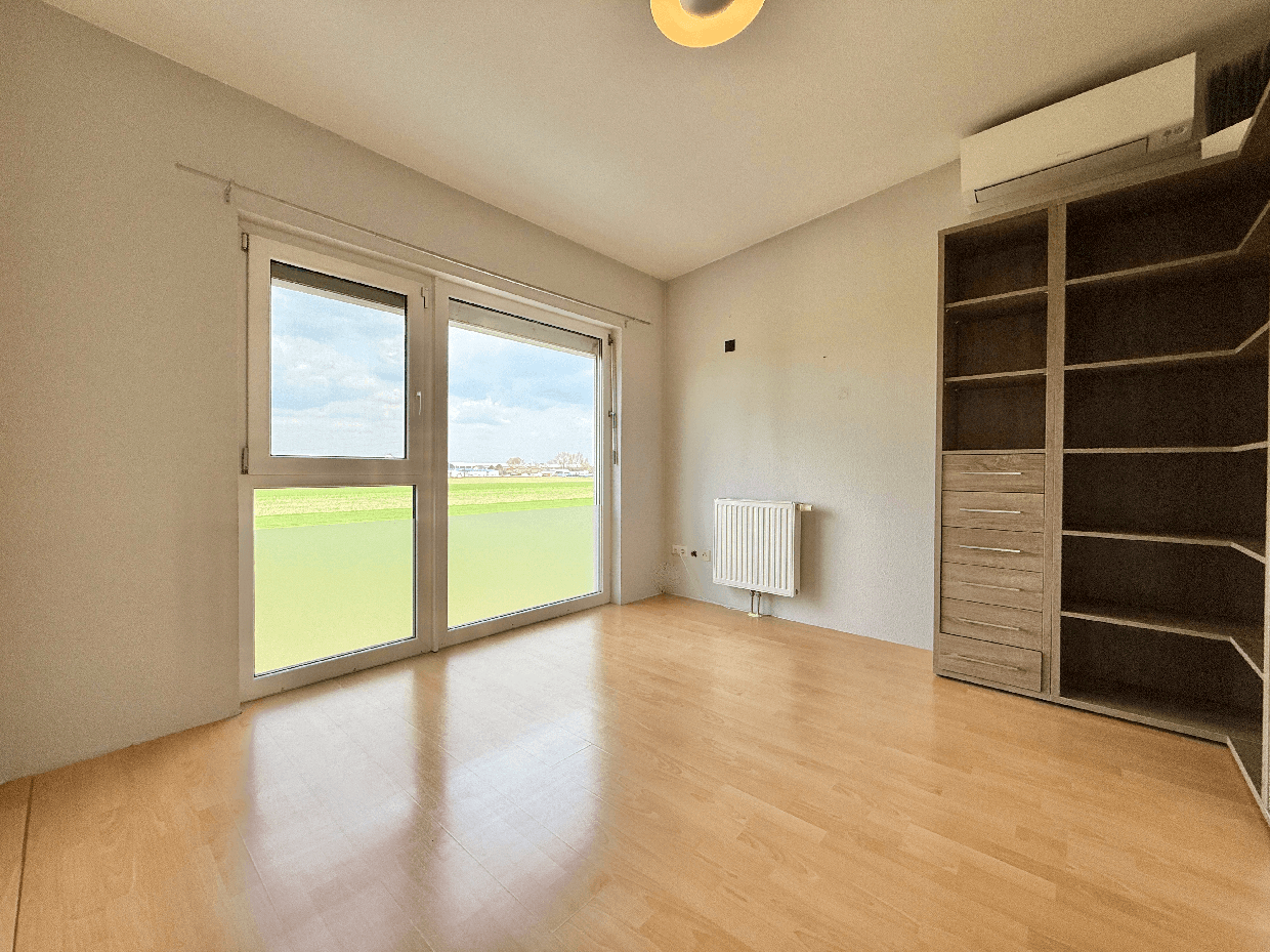 Leeres Schlafzimmer mit großem Fenster, Einbauschrank und Klimaanlage, bietet Blick ins Grüne.