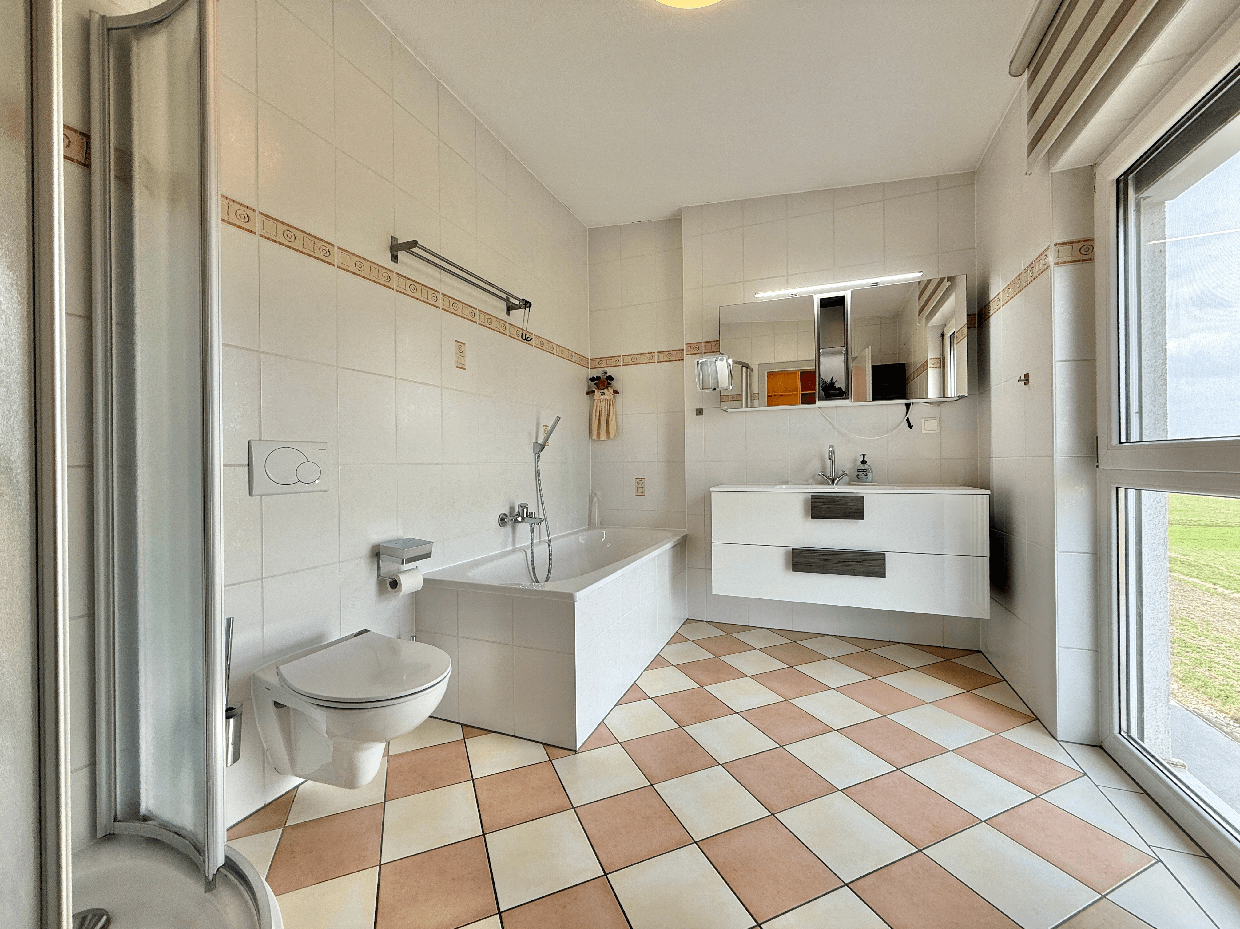 Badezimmer mit Badewanne, Dusche, WC und Fenster, ausgestattet mit hellen Fliesen und Spiegelschrank.