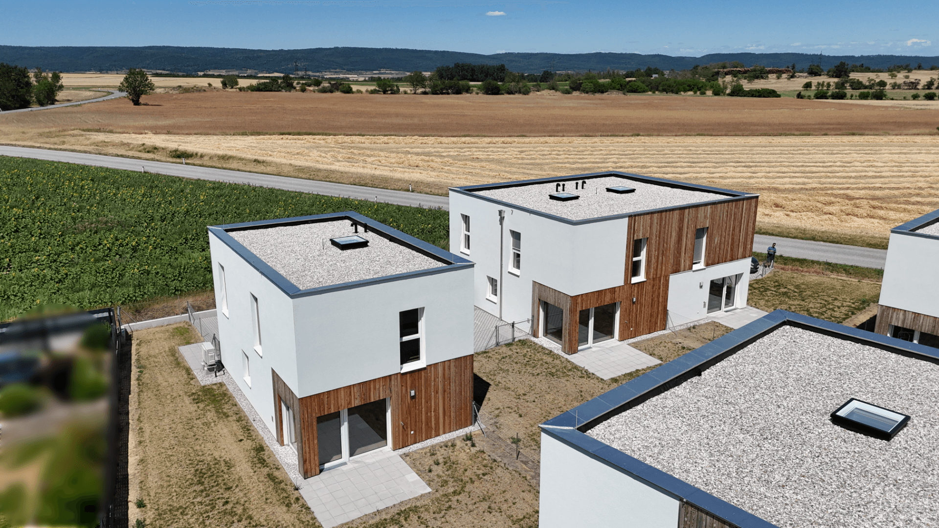 Luftaufnahme mehrerer moderner Häuser mit Flachdächern und angrenzenden Feldern unter blauem Himmel.