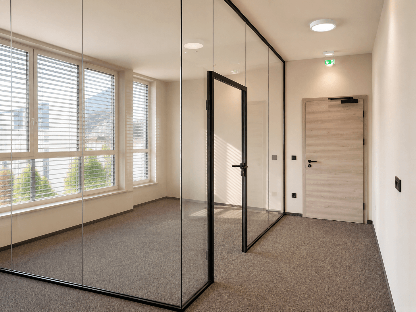 Modernes Büro mit Glaswänden, Teppichboden und großen Fenstern, die viel Tageslicht hereinlassen.