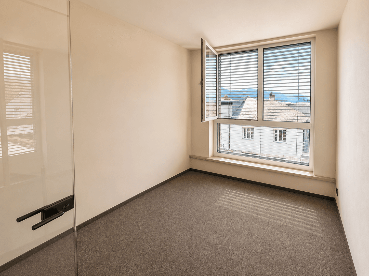 Leerer Büroraum mit Teppichboden und einem geöffneten Fenster, das frische Luft und Tageslicht hereinlässt.